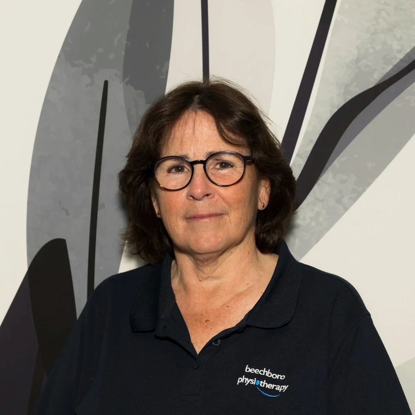 A woman with glasses and dark brown hair wearing a black polo shirt with the logo 'beechboro physiotherapy' printed on it, standing in front of an abstract background.