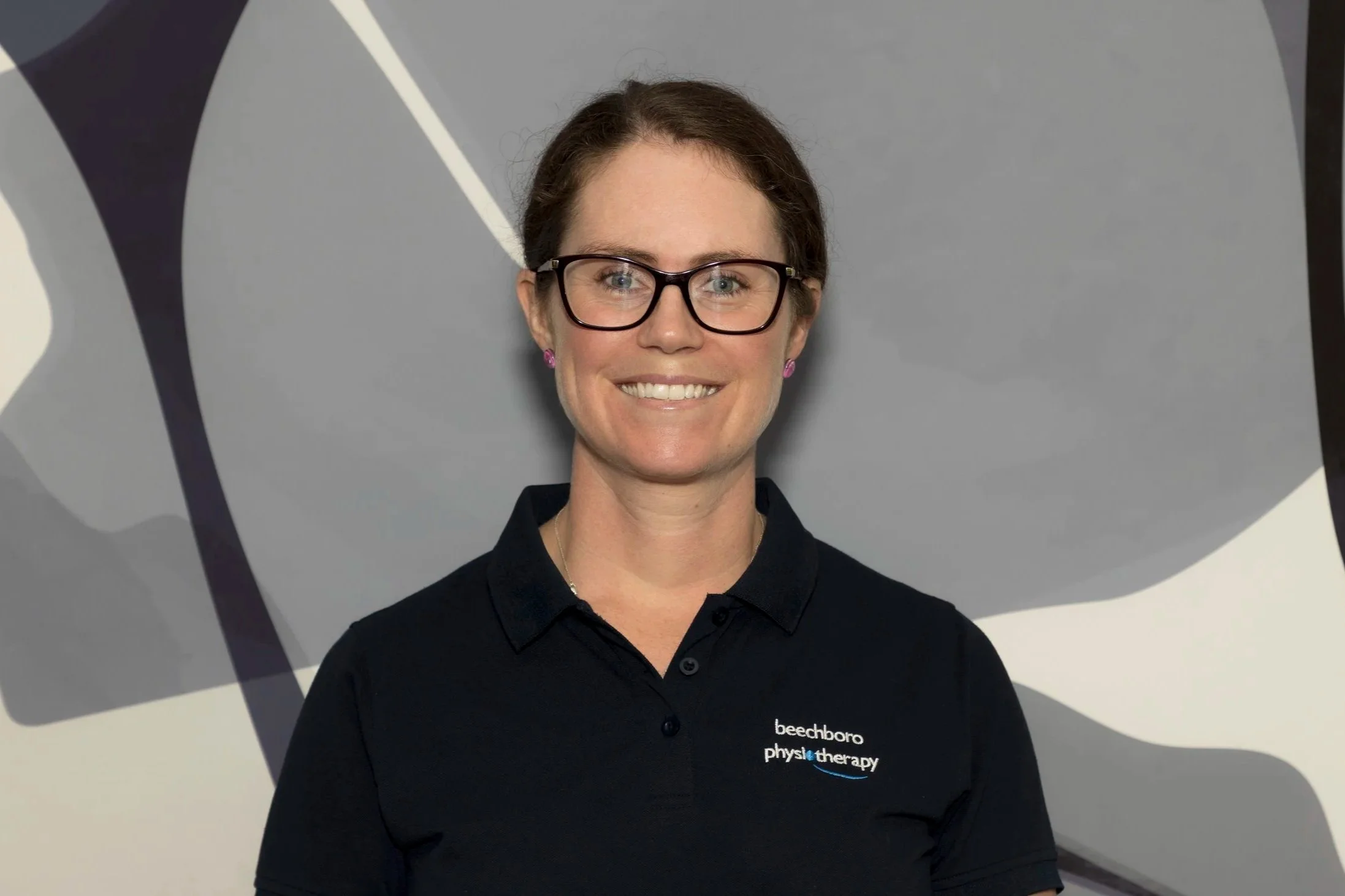 A woman with glasses and short brown hair smiling, wearing a black shirt with 'beechboro physiotherapy' logo, standing in front of a gray and white abstract background.
