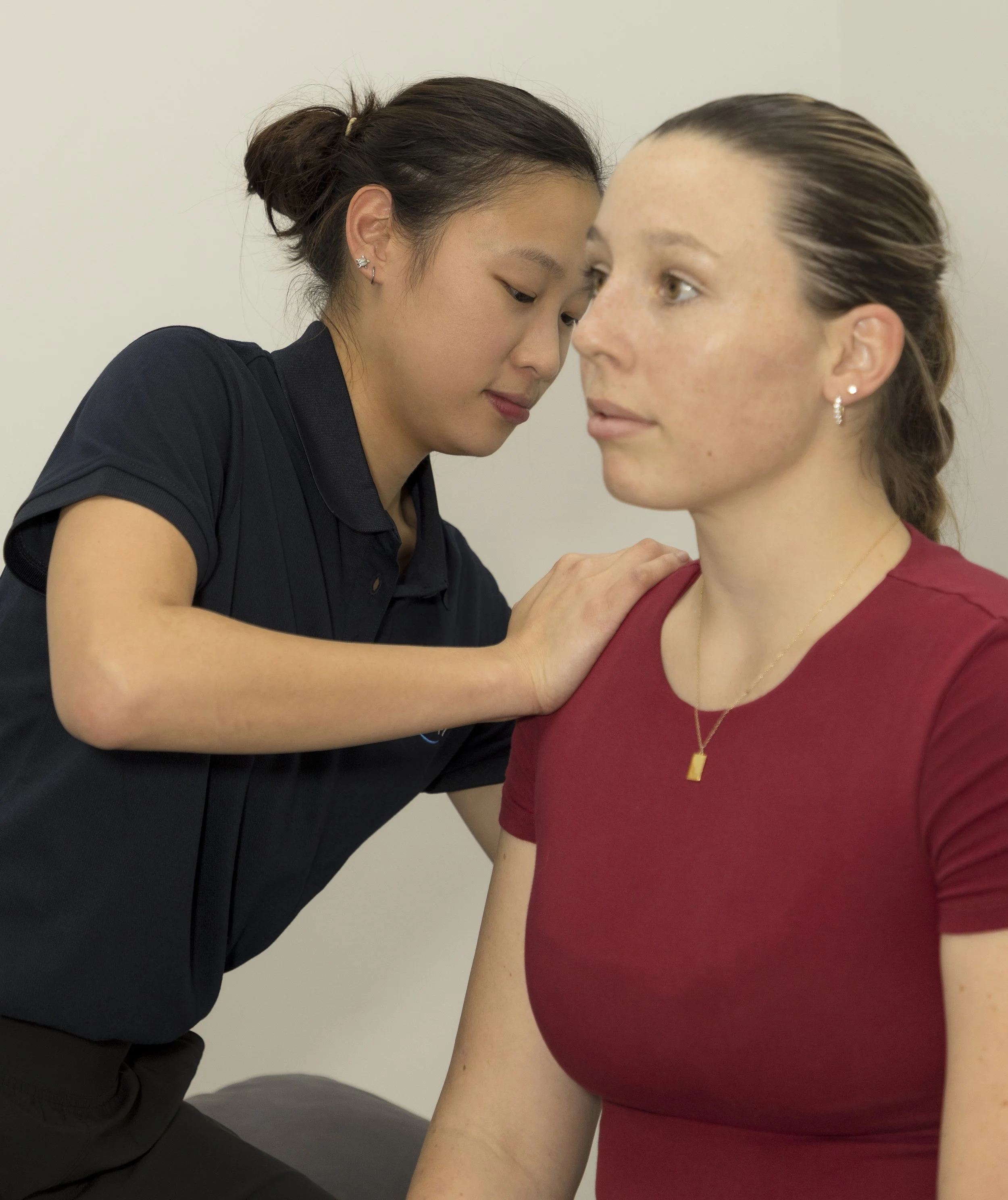 A healthcare professional performs a shoulder examination on a woman with fair skin and brown hair in a bun, wearing a red shirt and gold jewelry, in a clinical setting.
