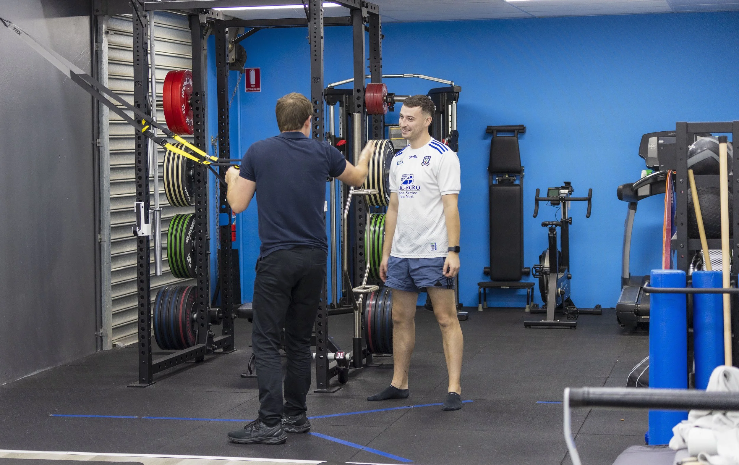 Physiotherapist and client in a gym, one in a navy shirt and the other in a white sports jersey, smiling and talking near a barbell with weight plates on a squat rack.