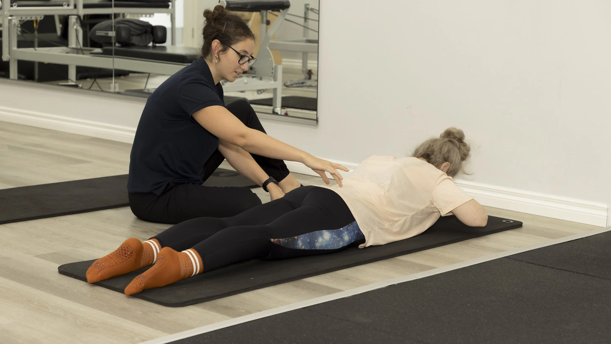 A Physiotherapist is helping a client perform a pilates pose on a black yoga mat indoors. The client is lying face down with her arms extended forward, and the Physiotherapist is assisting her.