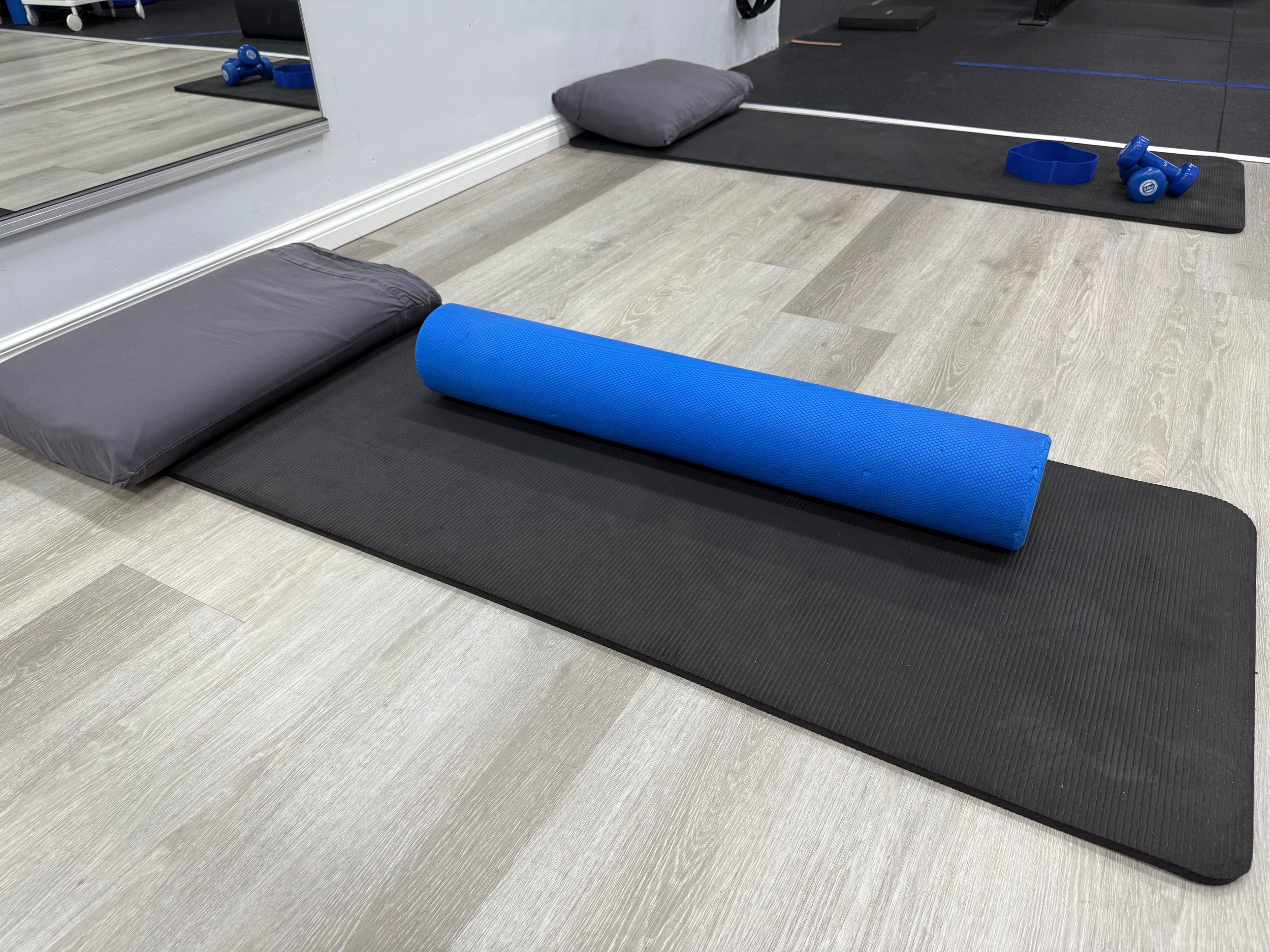 Exercise mats with a pillow, foam roller, and small dumbbells on a light wood floor, in a fitness or yoga studio.