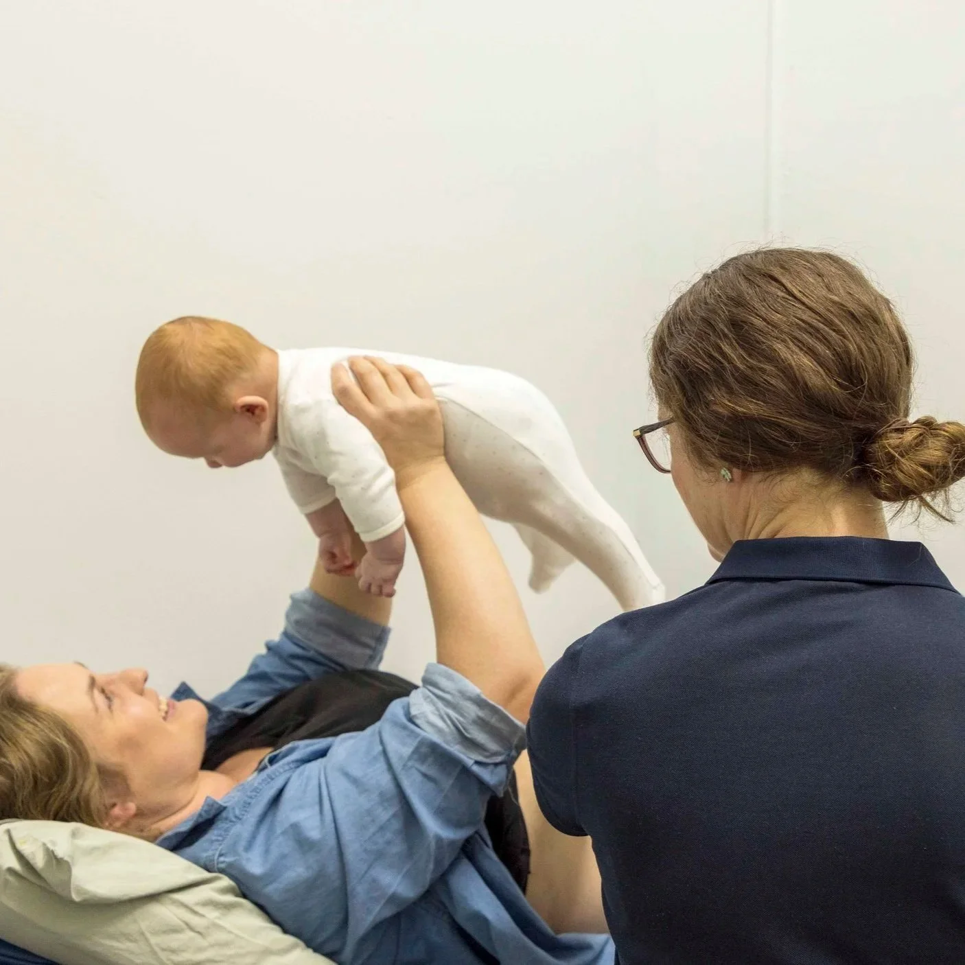 A woman lying on a bed holding a baby in the air, with a woman in glasses and a navy shirt assisting in the activity, in a room with plain walls.