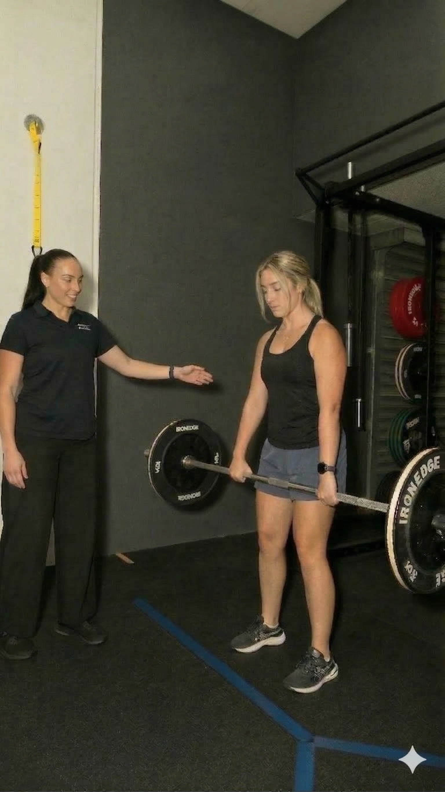 A woman performing a deadlift exercise with a barbell in a gym, supervised by a trainer.