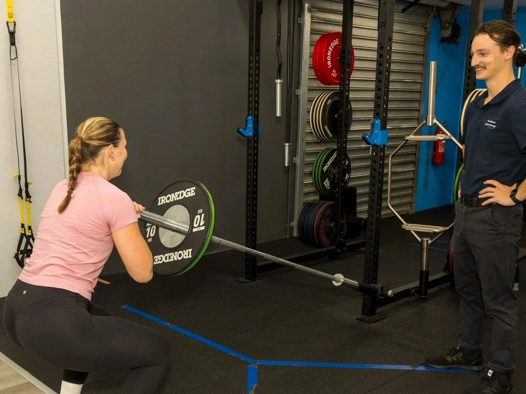 A woman performing a squat exercise with a barbell and weight plates while a man watches and supervises in a gym.