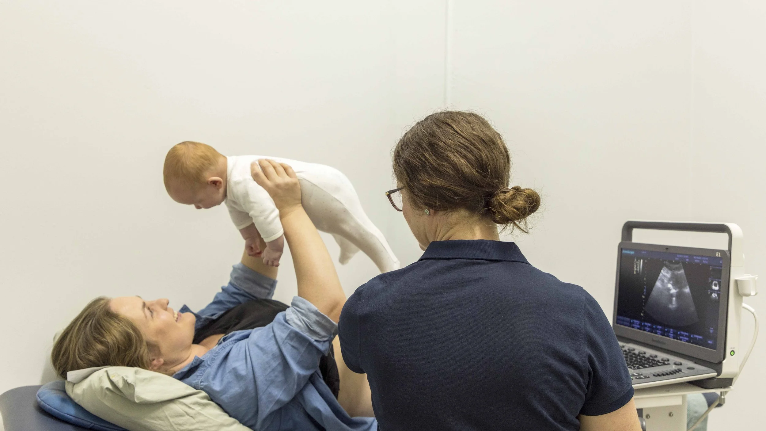 A woman lying on a medical examination table holding a baby above her, while a healthcare professional watches and an ultrasound monitor displays a sonogram.