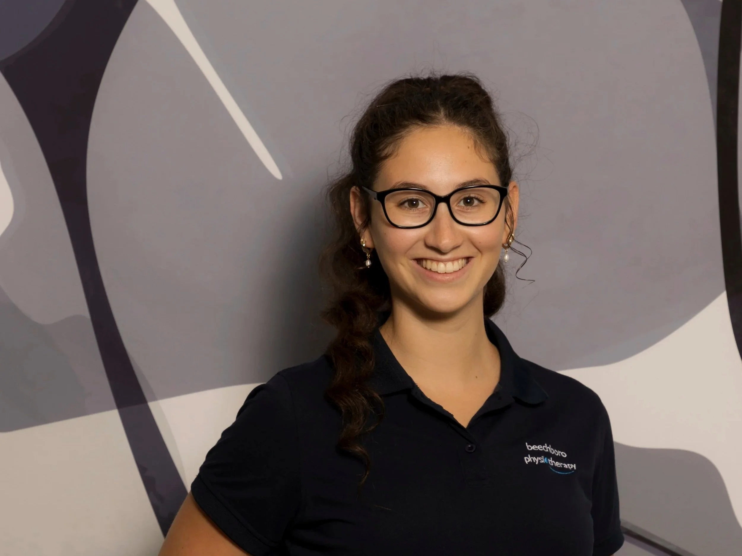 A smiling woman with glasses and earrings wearing a navy polo shirt with a logo that reads 'beachdune physiotherapy' in front of an abstract gray and white background.