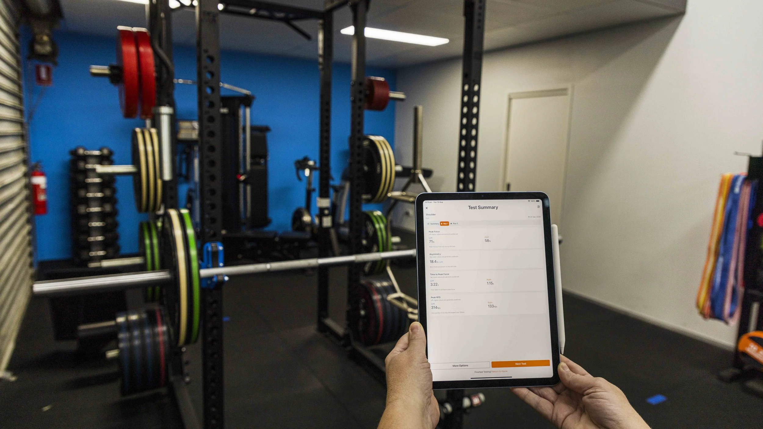 Person holding a tablet in front of a gym weightlifting area with loaded weight plates on black racks.