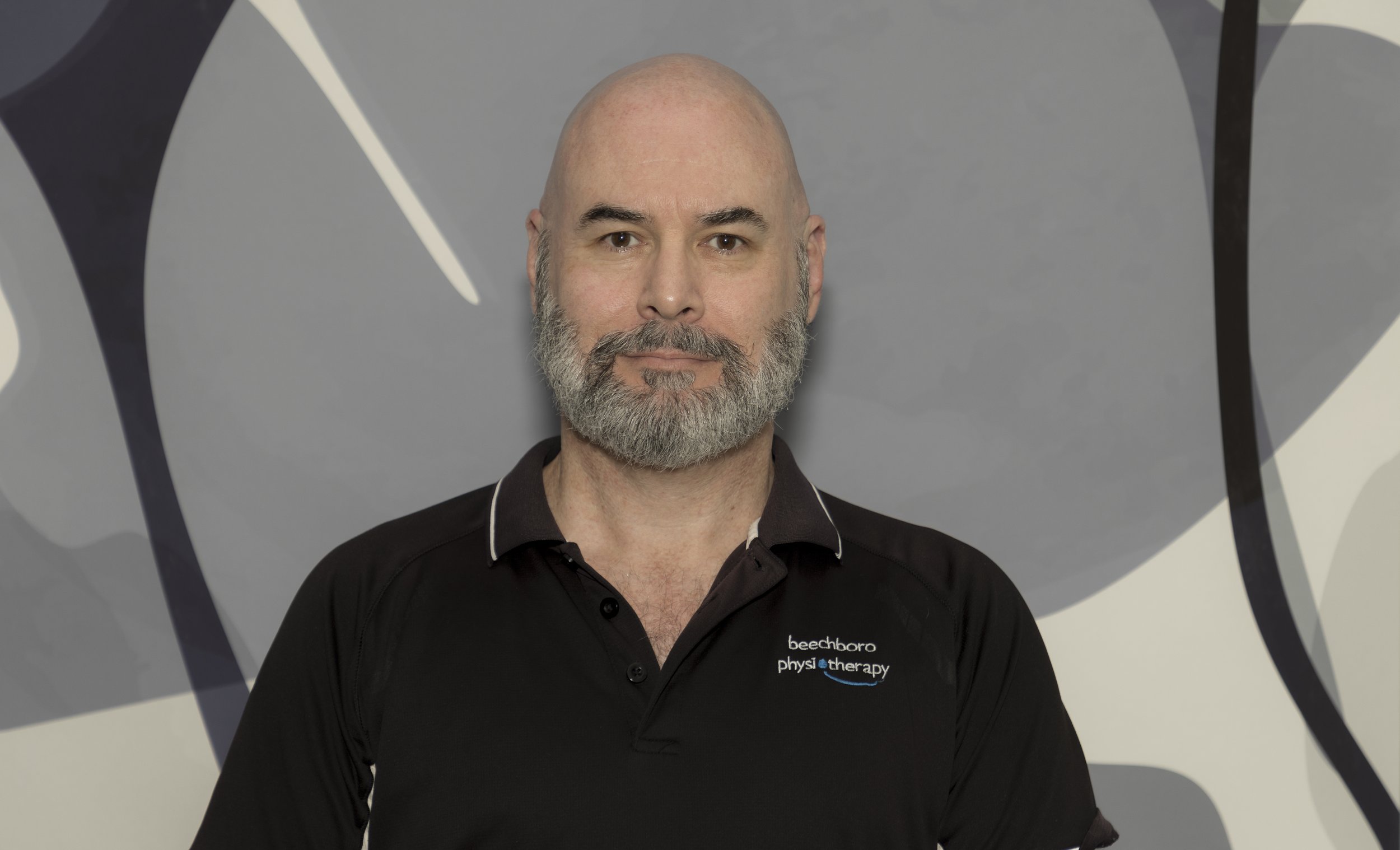 Man with a bald head and gray beard wearing a black polo shirt with white and blue logo, standing in front of an abstract black, white, and gray background.