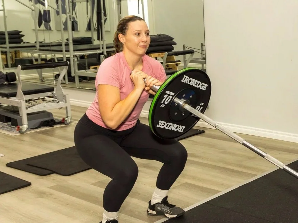 A woman in a pink shirt doing a squat with a barbell in a gym.