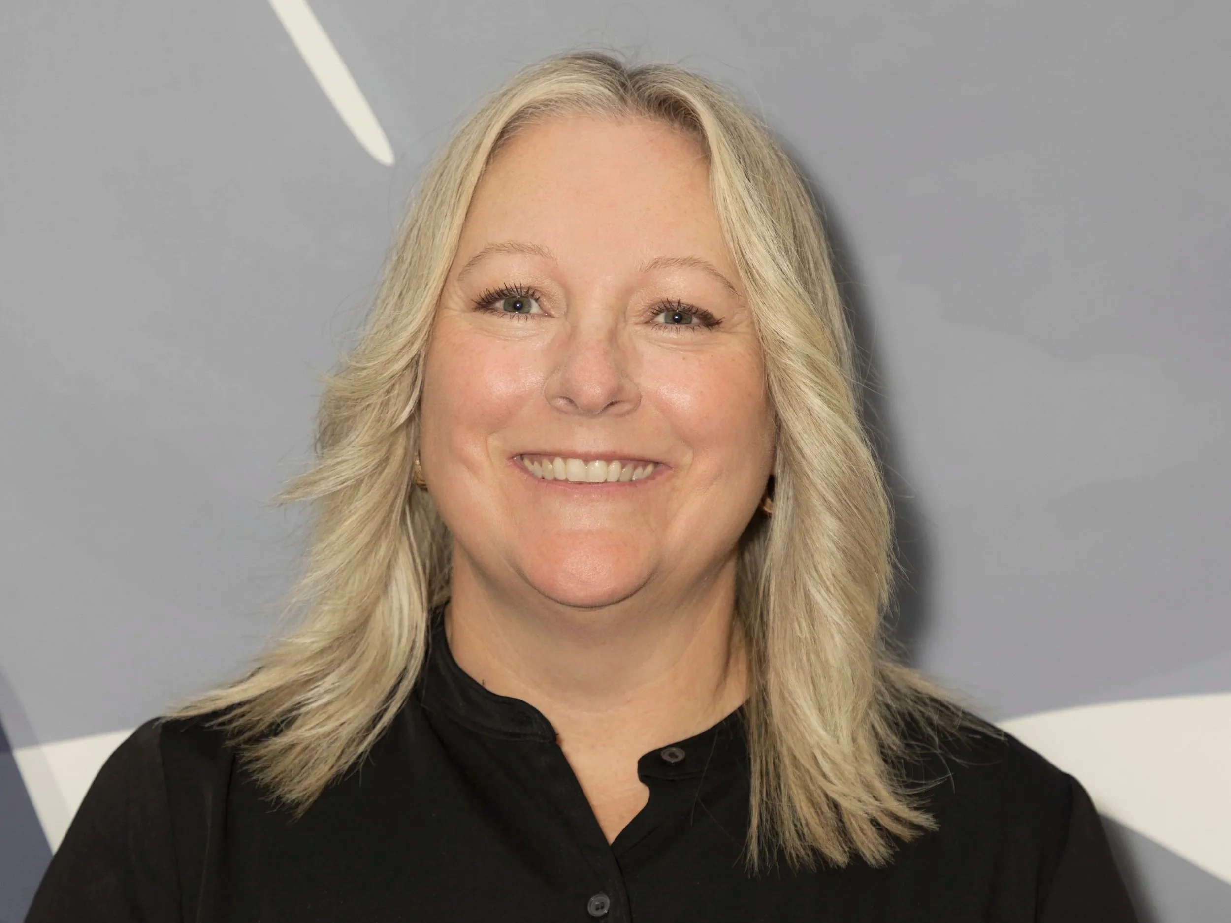 Tracey Scanlan with blonde hair smiling, wearing a black top, standing in front of a gray background with abstract white and gray shapes.