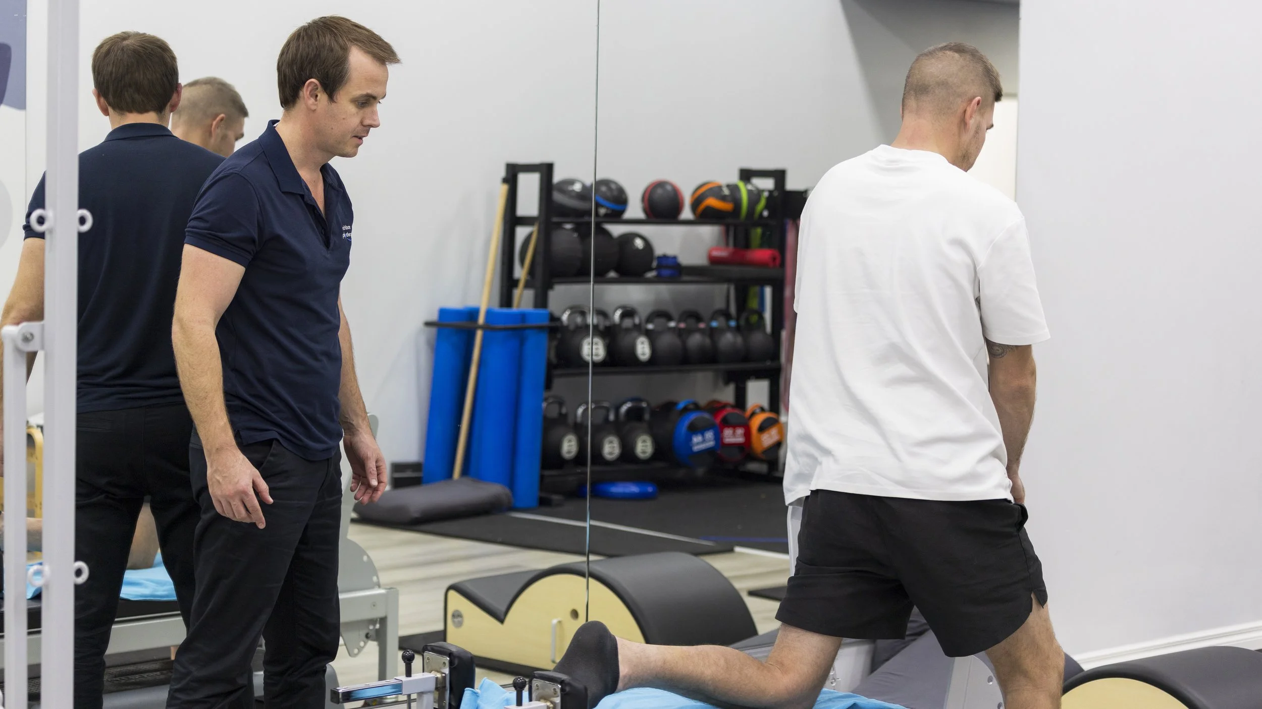 Physiotherapist assists a man stretching on a Pilates reformer iat Beechboro Physio with workout equipment on shelves in the background.