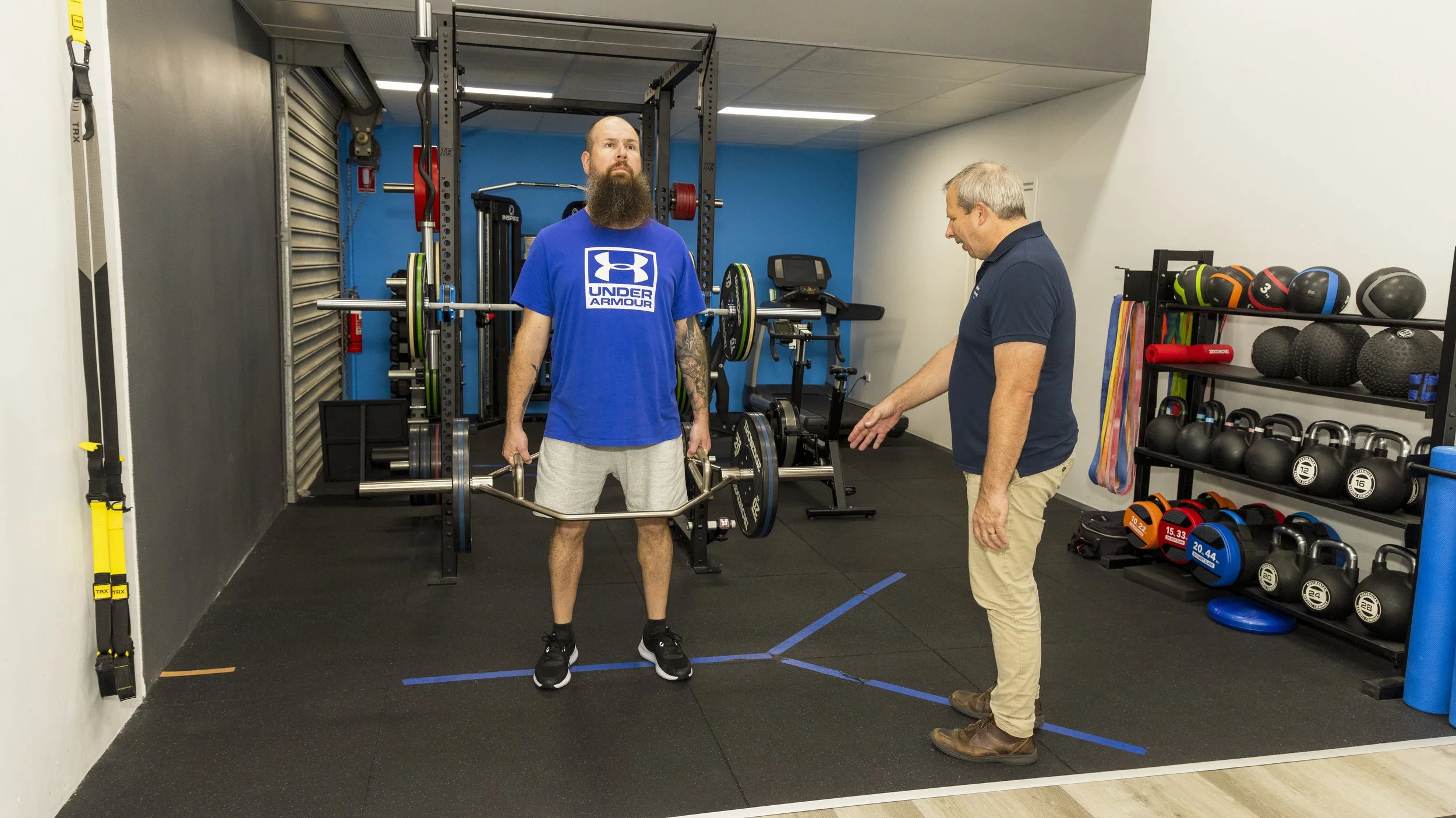 A man with a beard and tattooed arm is lifting a barbell in a gym while a trainer explains something to him. The gym has weight equipment, colorful medicine balls, and resistance bands.