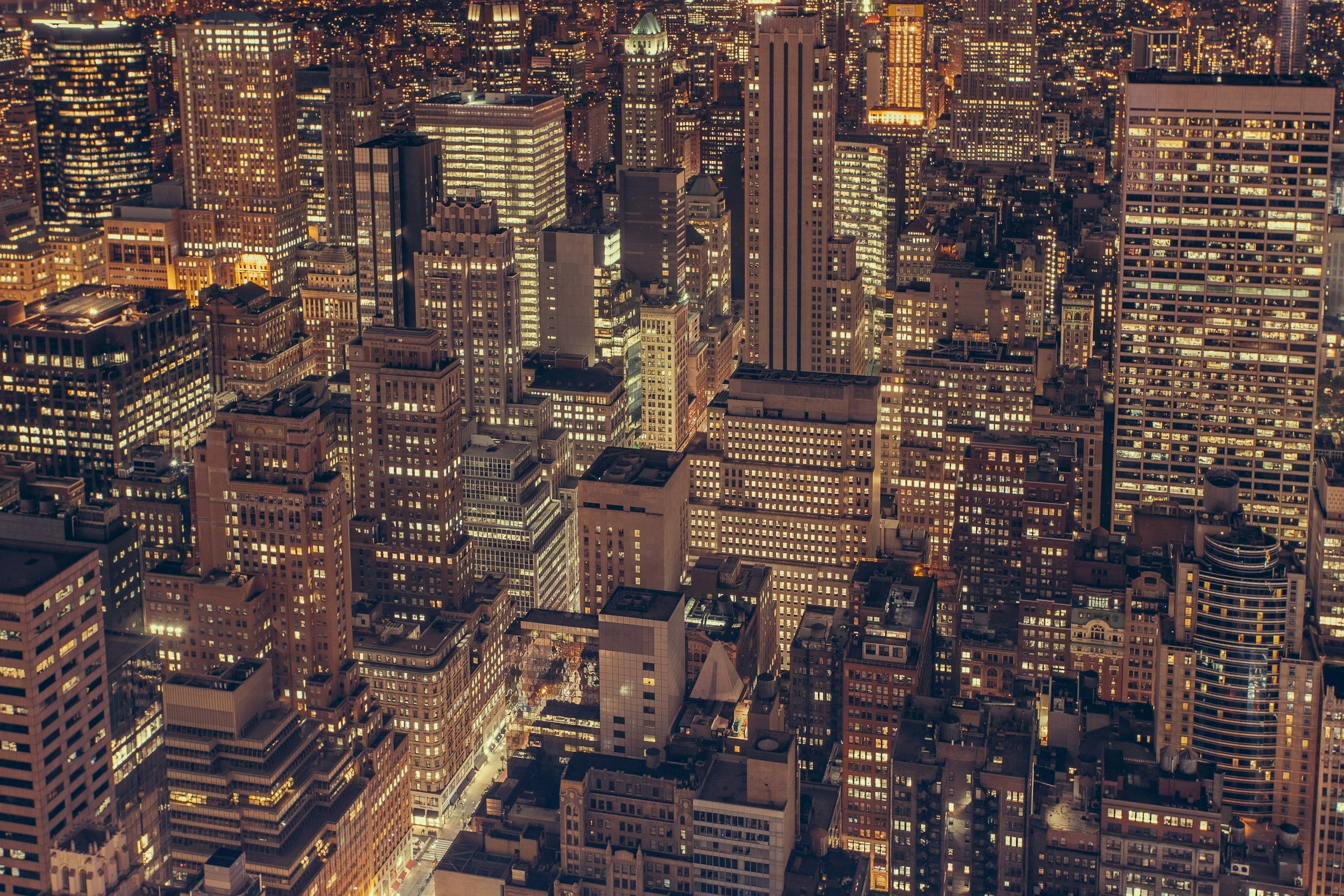 A nighttime aerial view of a dense cityscape with numerous illuminated skyscrapers and high-rise buildings.
