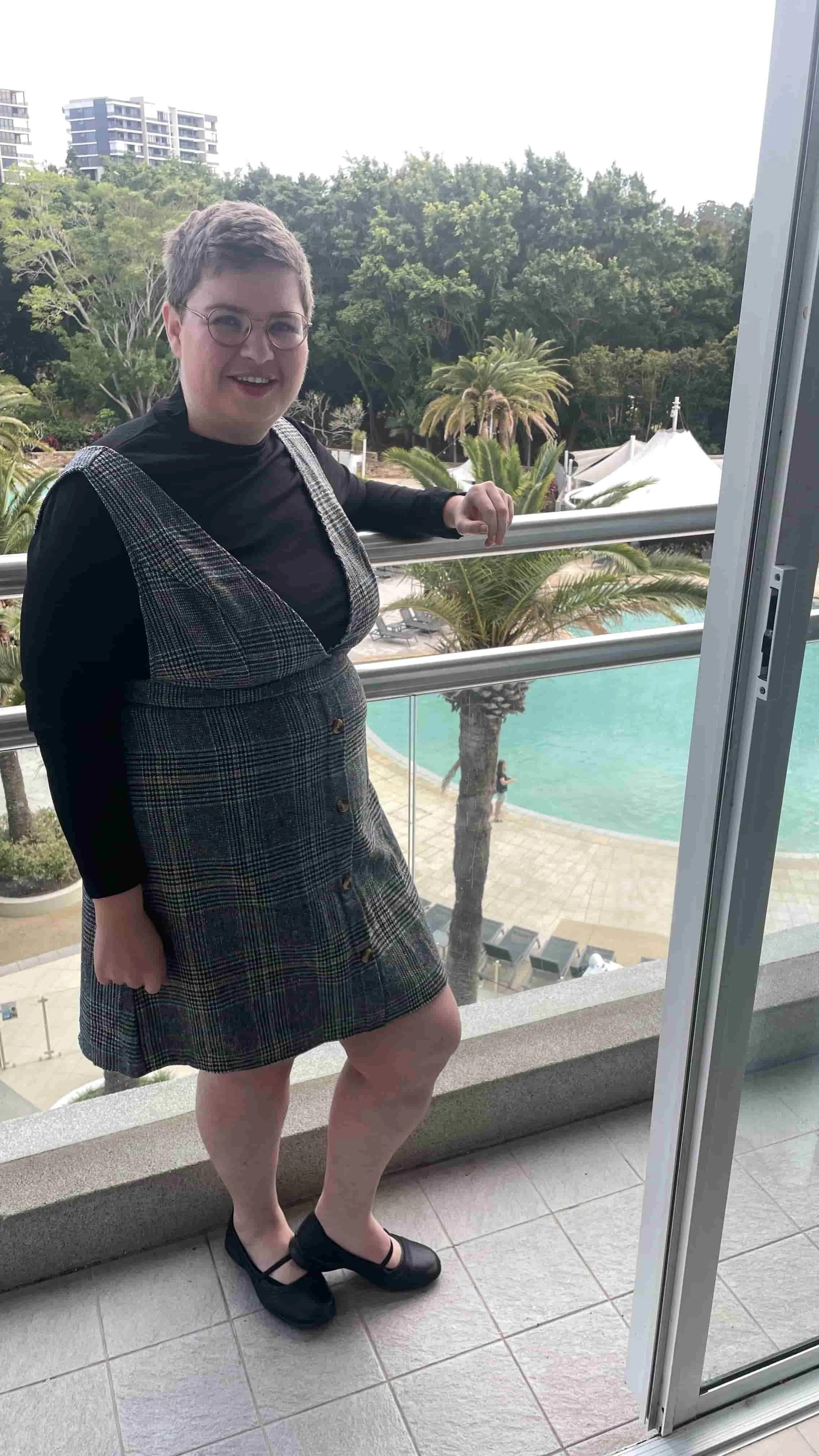 Karen from Karen's Job Services, in glasses and a plaid dress, stands on a balcony overlooking a pool and palm trees, smiling at the camera.