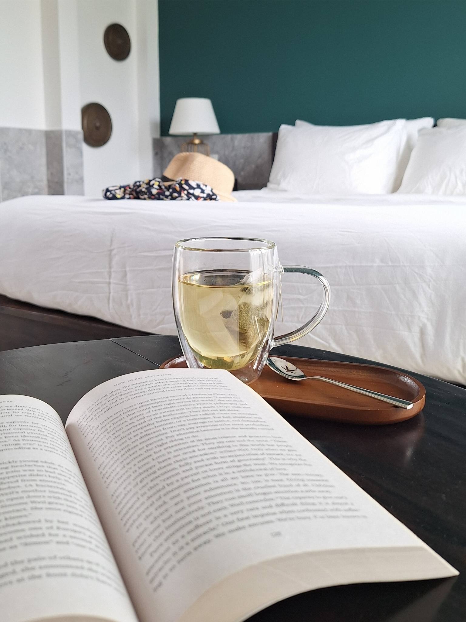 Open book and glass of herbal tea on a dark wood table with a white-linen bed and teal wall in the background