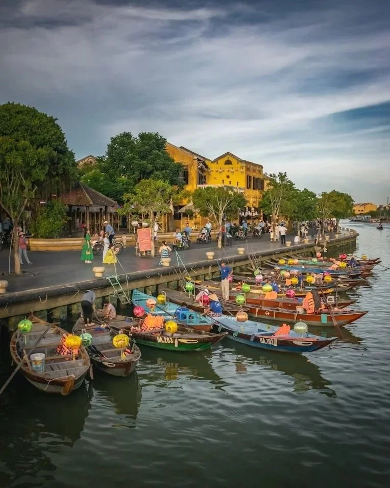 Boats docked along a riverwalk with people strolling and engaging in activities in a vibrant, lively setting.