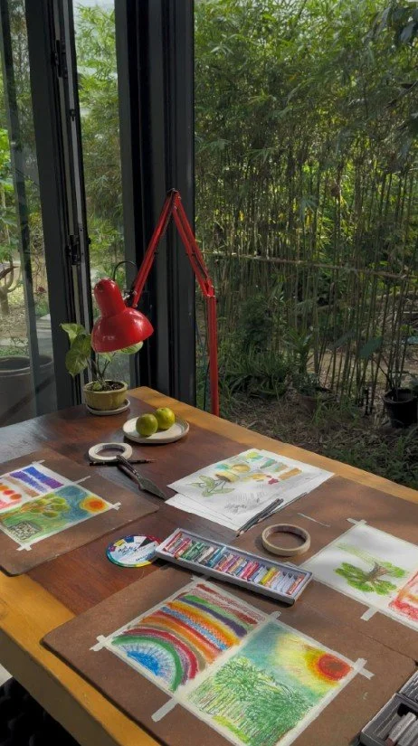 Watercolor and oil pastel paintings spread on a studio table with paints, a red desk lamp, and bamboo garden view