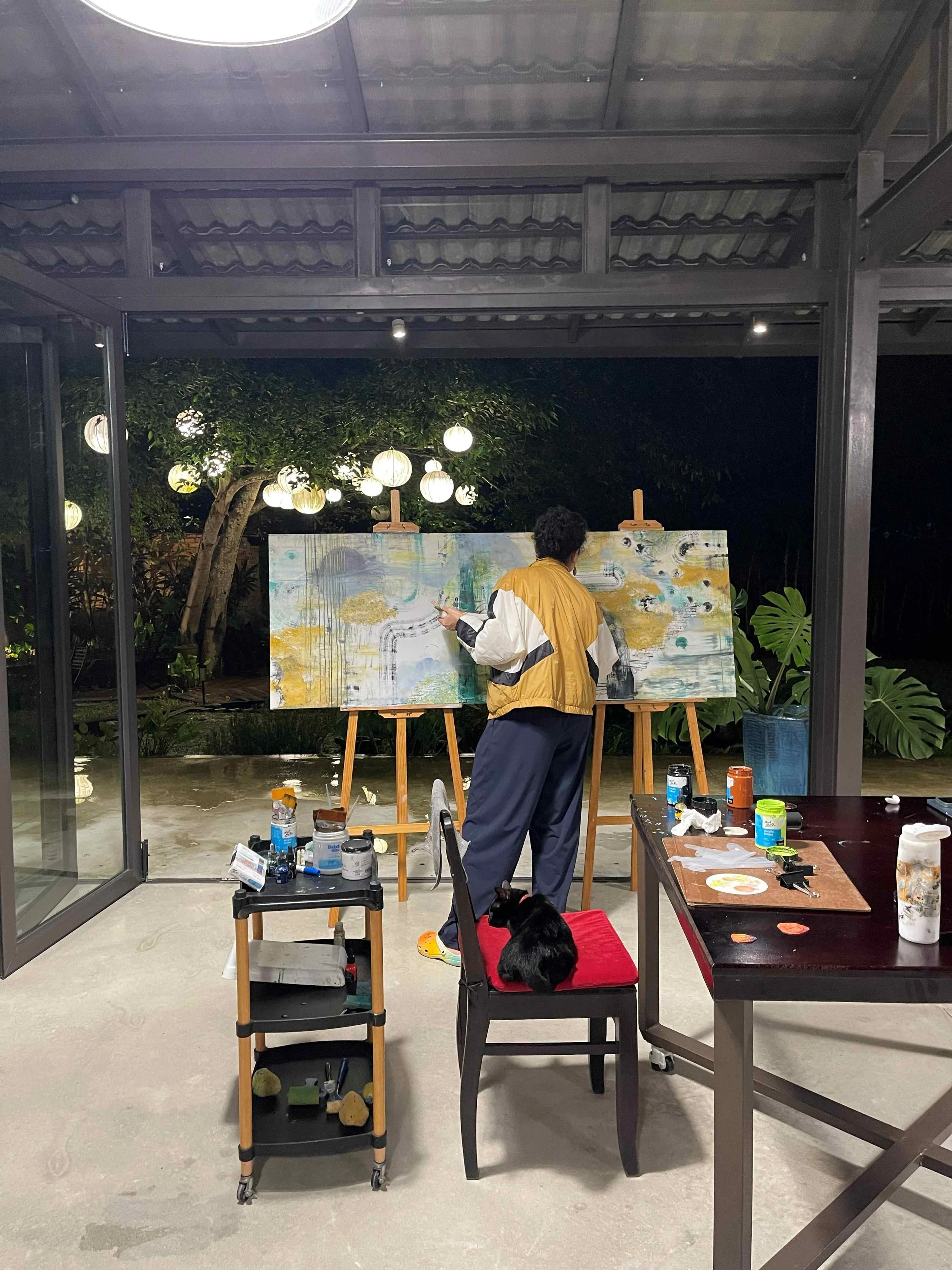 Artist painting a large abstract canvas at an easel in the studio at night with garden lanterns glowing