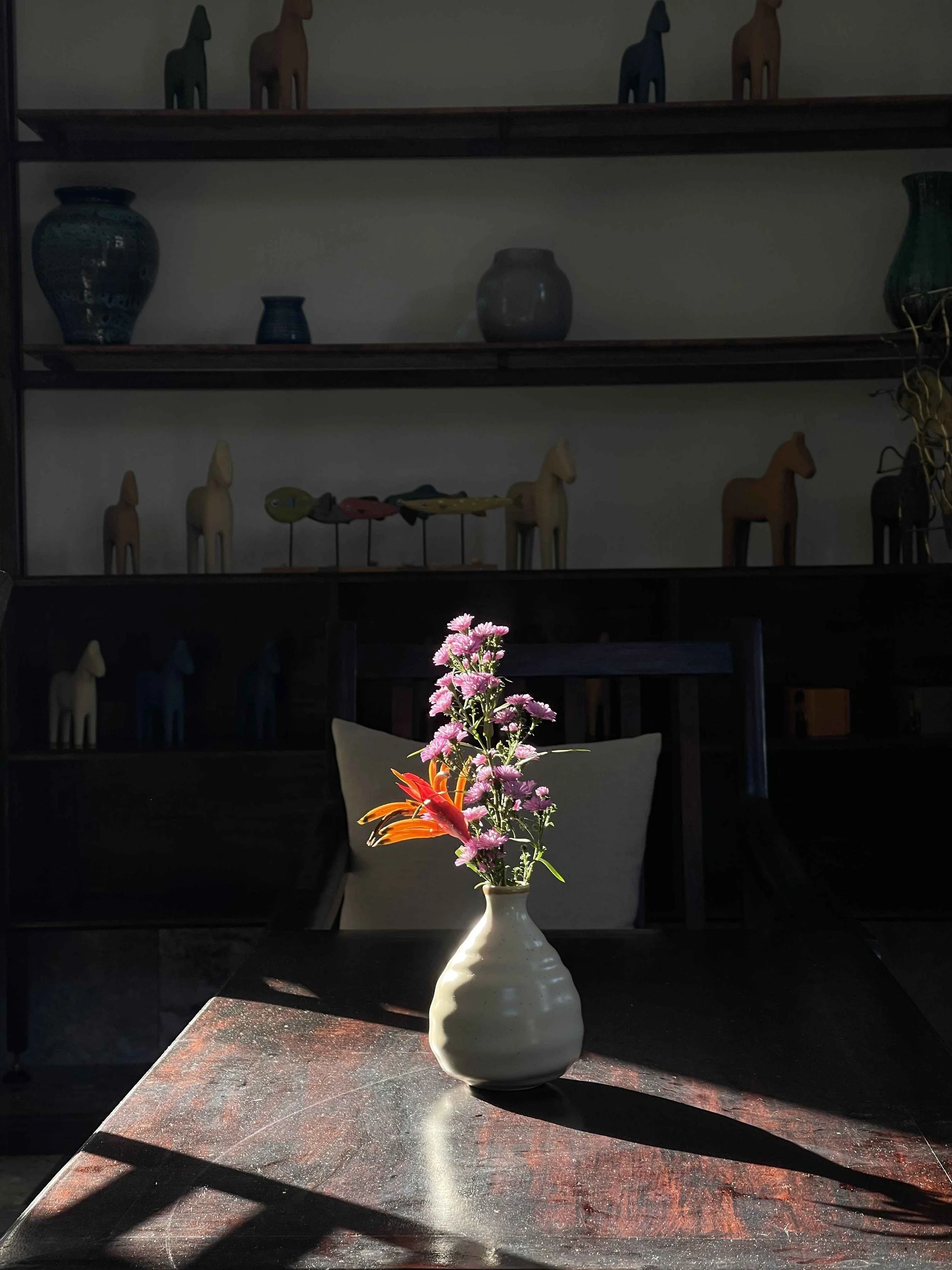 A white ceramic vase with pink and orange flowers on a dark granite table, illuminated by sunlight, with a dark background featuring shelves displaying decorative pottery and small horse figurines.