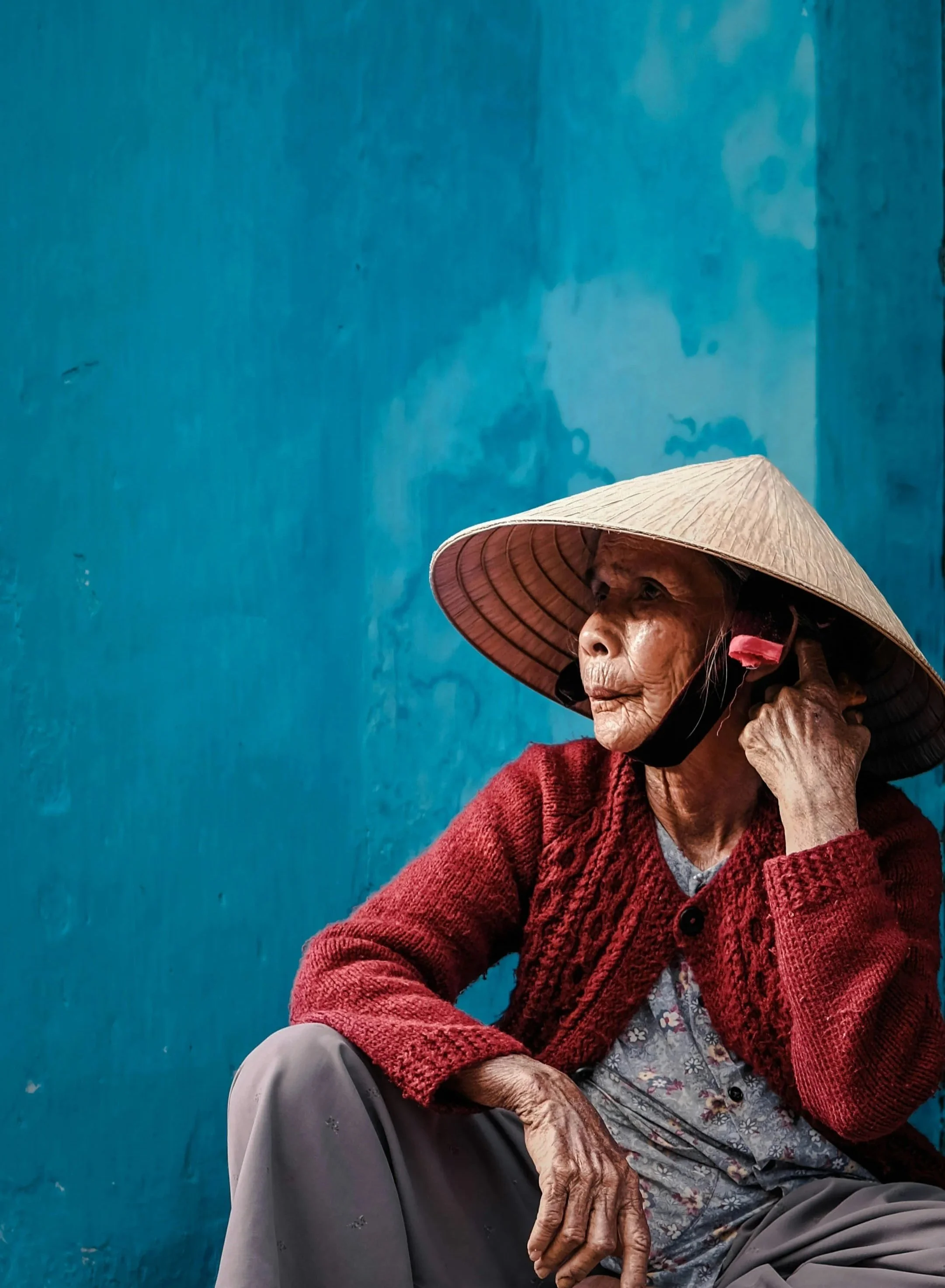 Elderly Vietnamese woman in a conical hat and red cardigan sitting against a blue wall portrait