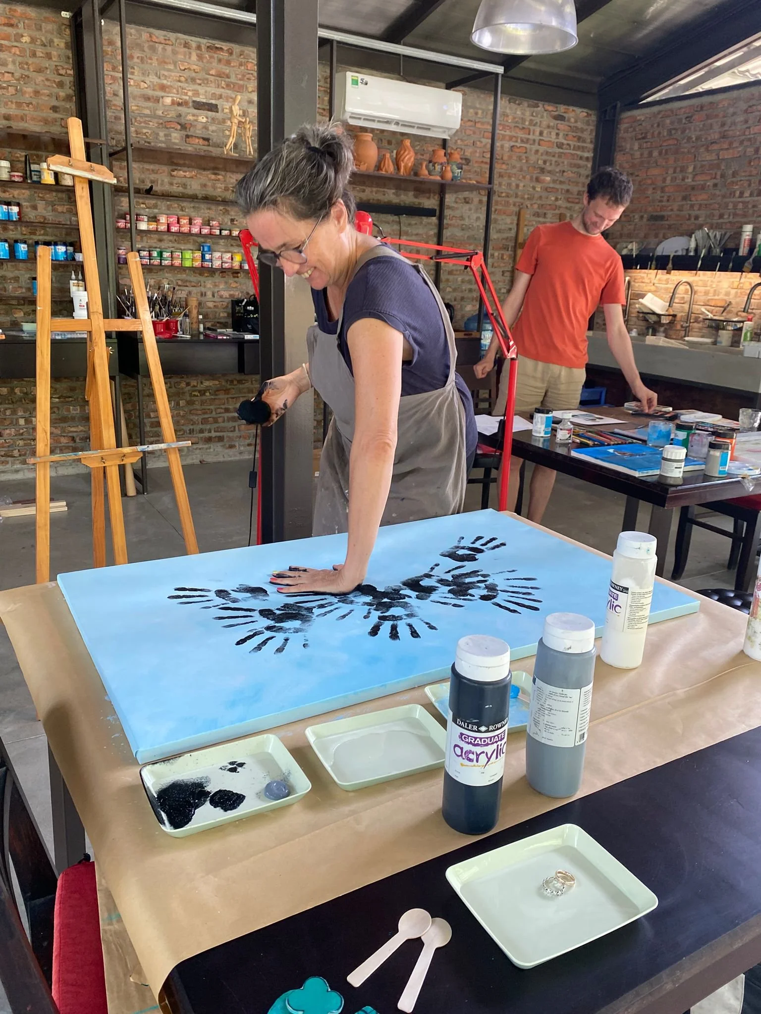 two guests in art studio working on big canvases with paint tubes and art supplies on tables
