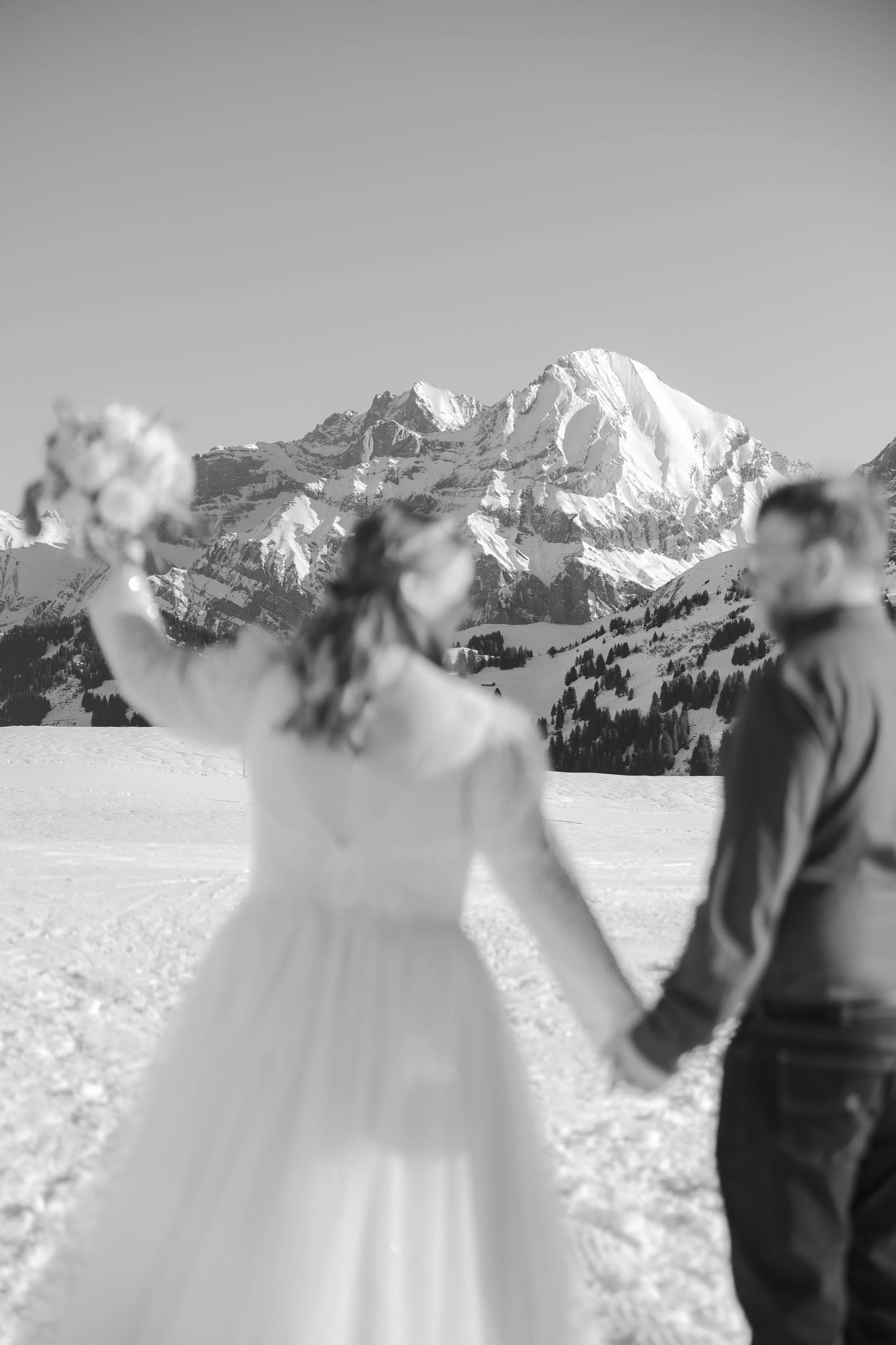 Verliebtes Paar in Hochzeitskleidung hält sich in verschneiter Berglandschaft an den Händen, im Hintergrund schneebedeckte Berge.