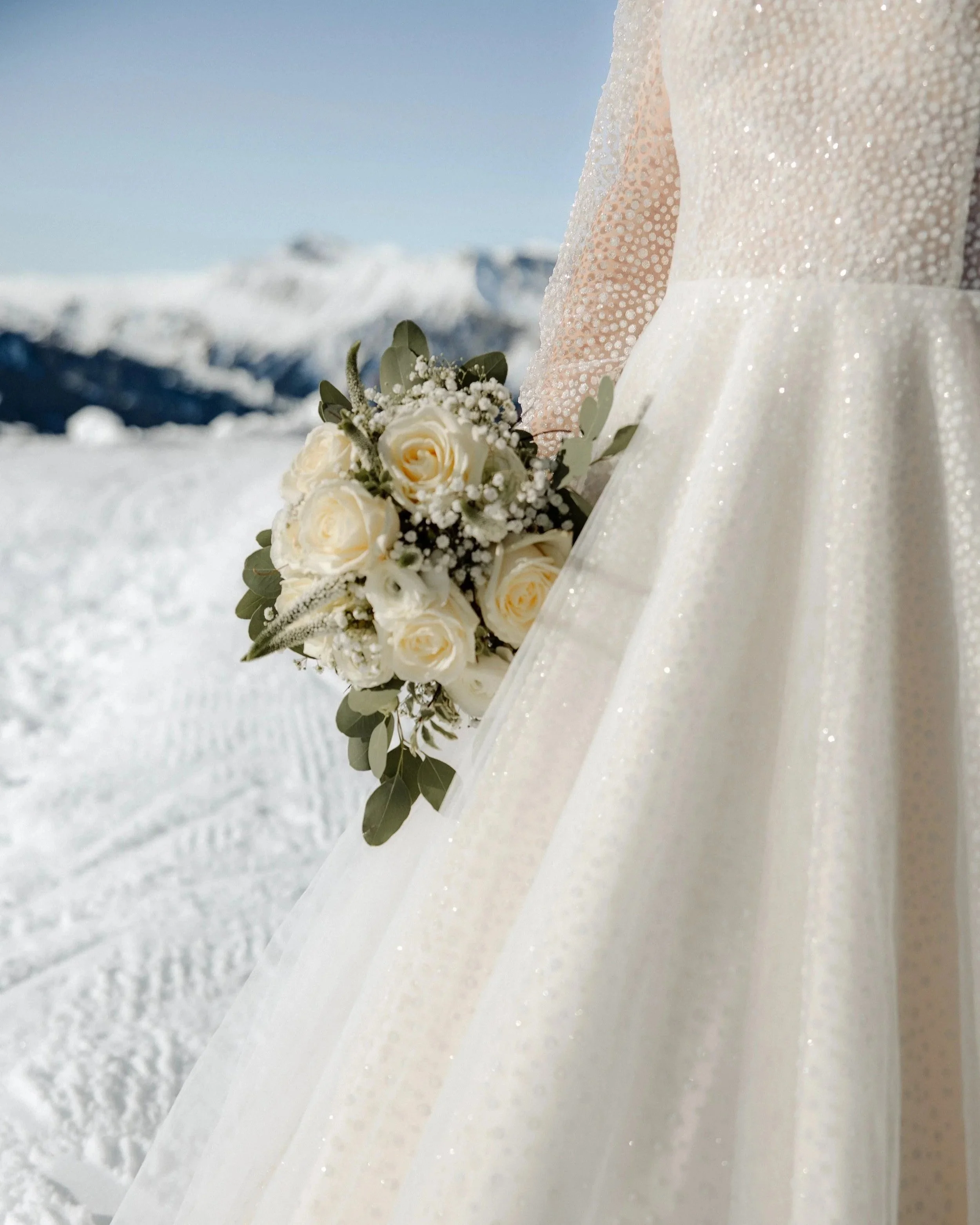 Brautkleid und Brautstrauß vor verschneiter Bergkulisse.