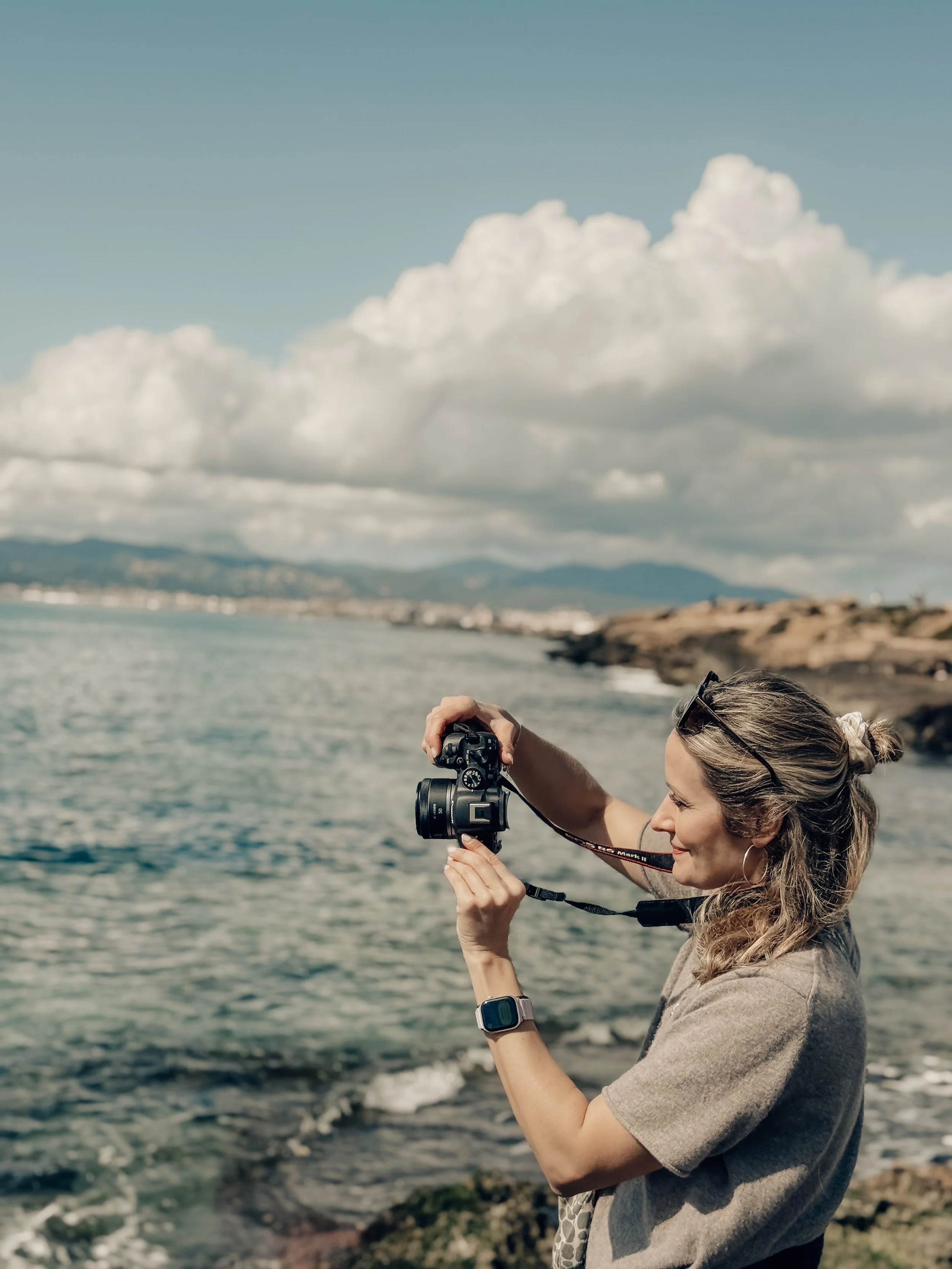 Frau fotografiert am Meer bei bewölktem Himmel mit Kamera, trägt Smartwatch, Sonnenbrille auf Kopf.