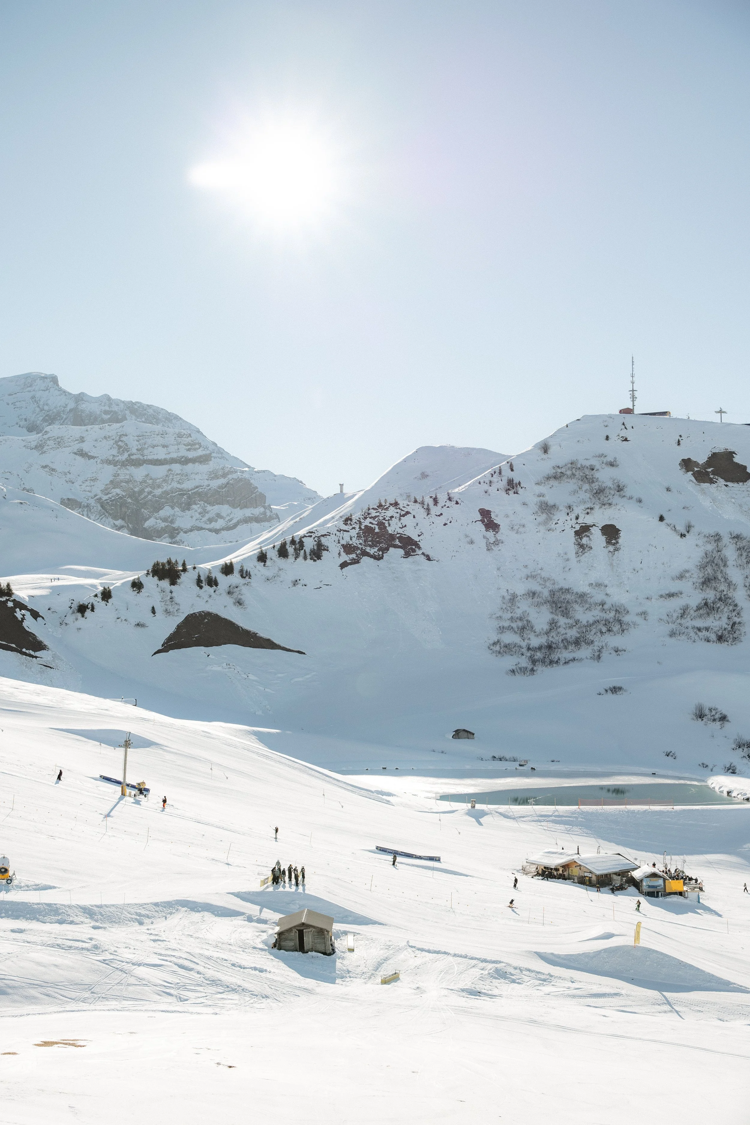 Schneebedeckte Berge und Hänge mit Skifahrern, Skiliften und kleinen Gebäuden unter einem sonnigen Himmel.