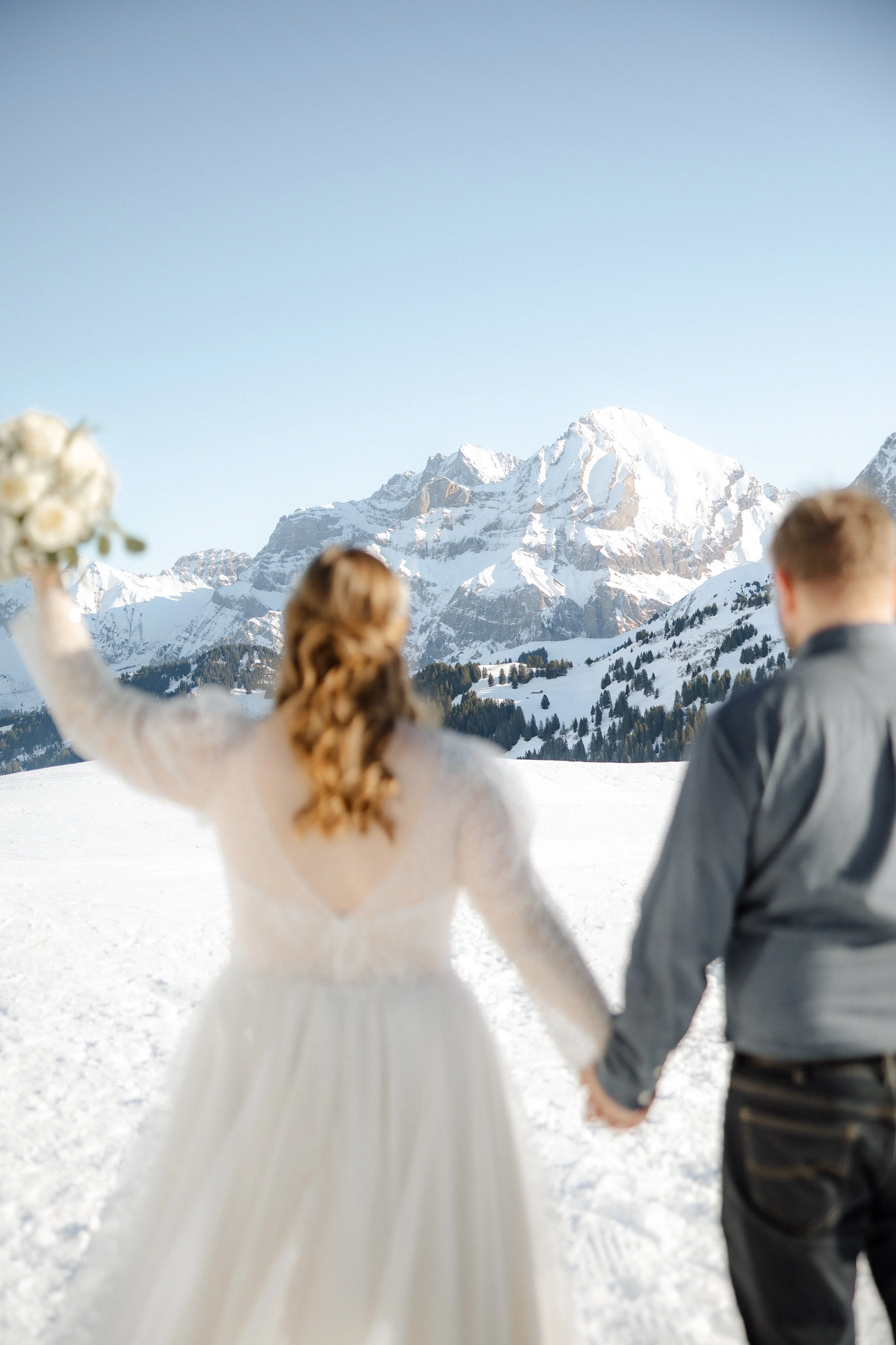 Ein Brautpaar hält Händchen in einer verschneiten Berglandschaft mit schneebedeckten Gipfeln im Hintergrund.