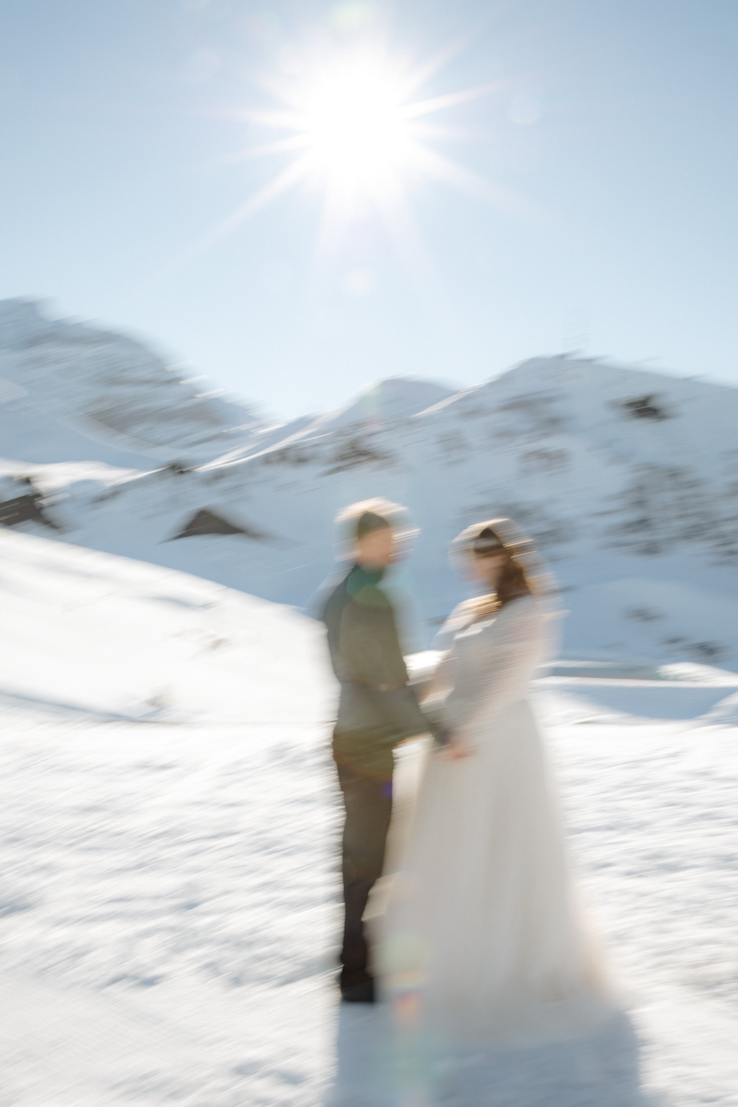 Verwackeltes Bild von einem Paar in formeller Kleidung, wahrscheinlich bei einer Hochzeitszeremonie, in einer verschneiten Berglandschaft bei Sonnenlicht.