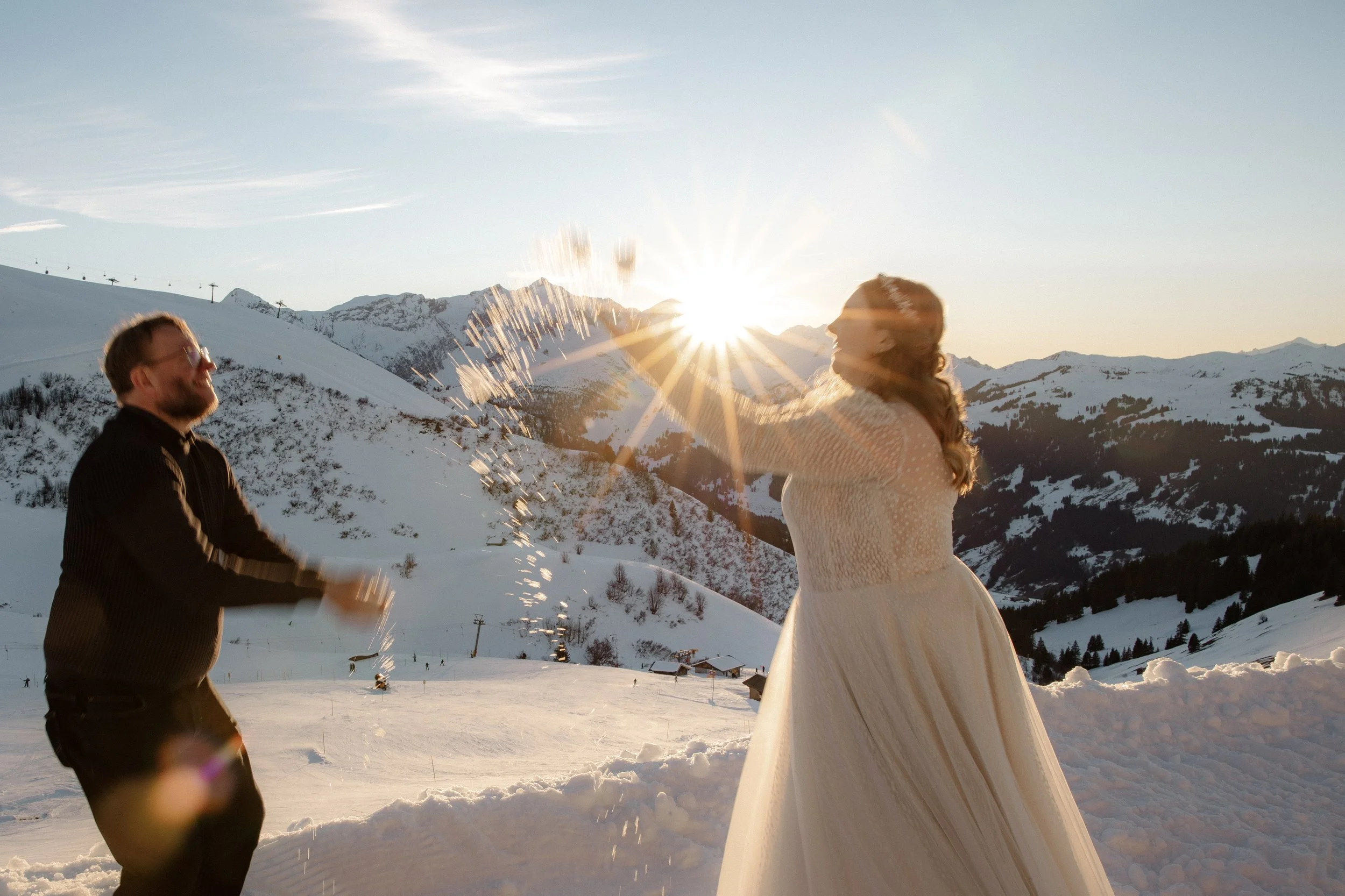 Ein Paar in formeller Kleidung spielt im Schnee in den Bergen mit Schneebällen bei Sonnenuntergang.