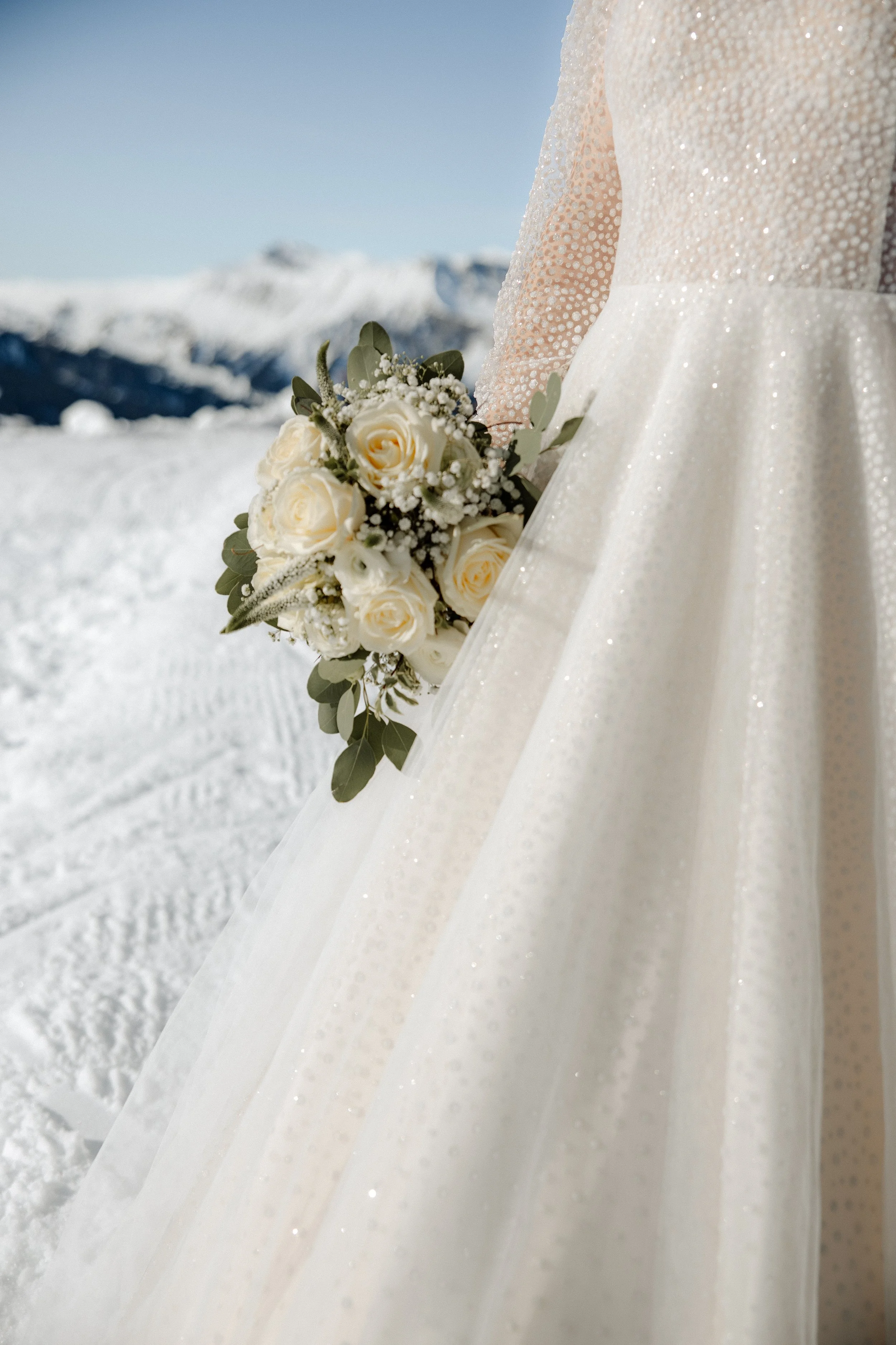 Nahaufnahme eines weißen Brautkleides und eines Brautstraußes mit weißen Rosen vor einer verschneiten Bergkulisse.