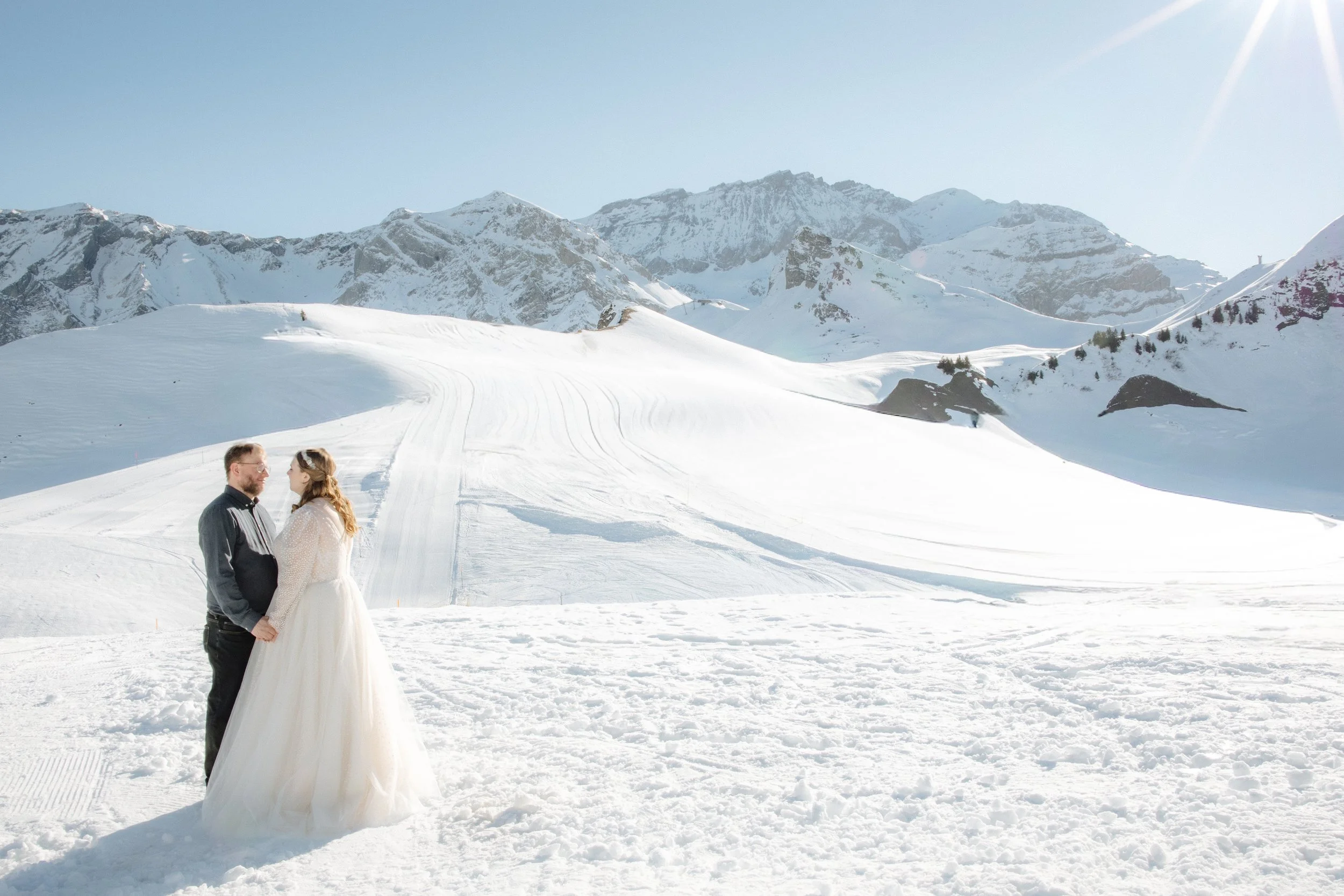 Ein Paar steht sich in der verschneiten Landschaft der Alpen gegenüber, die Frau trägt ein weißes Kleid, der Mann eine dunkle Jacke, im Hintergrund schneebedeckte Berge und strahlend blauer Himmel.