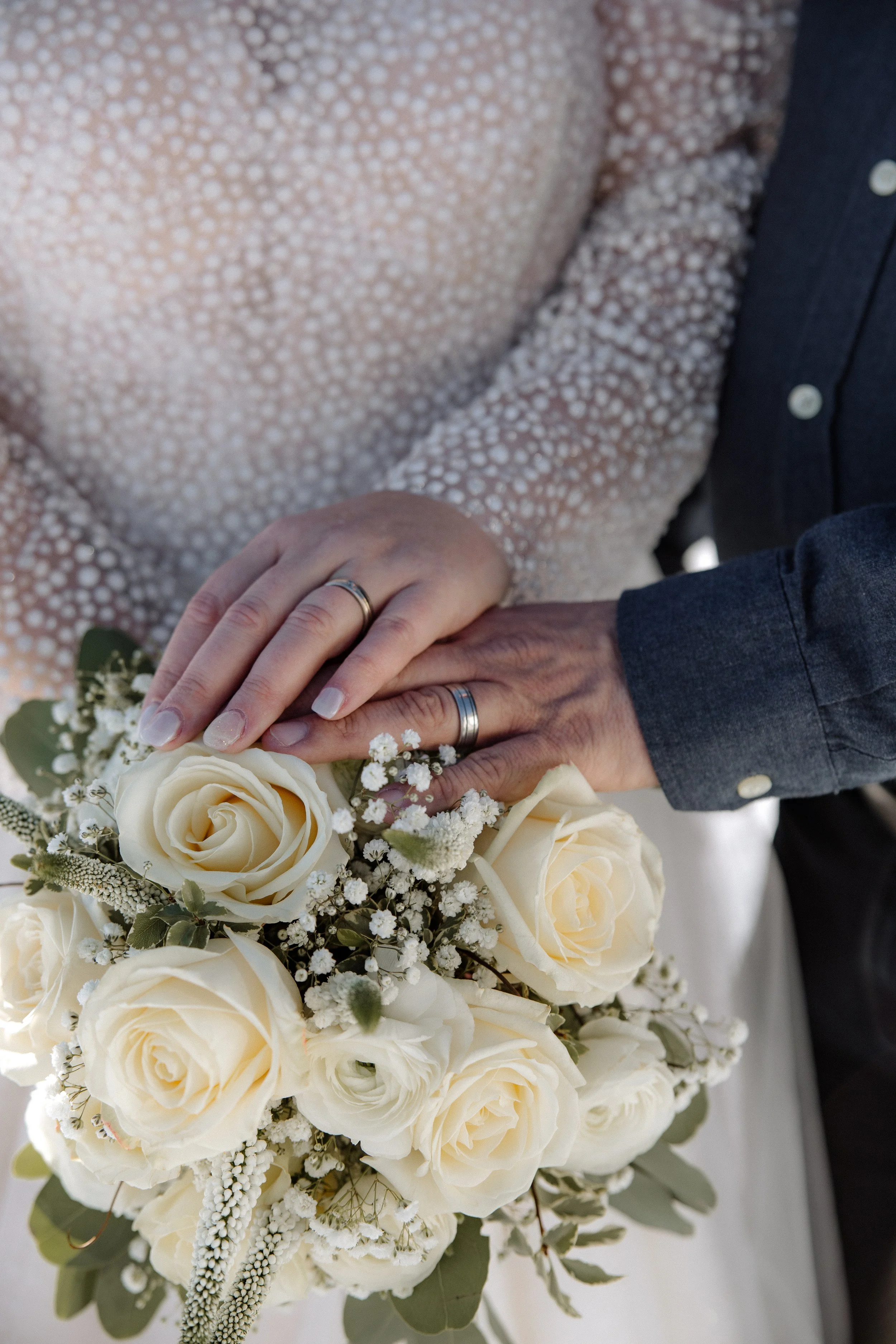 Hände eines Brautpaares mit Eheringen auf einem weißen Blumenstrauß aus Rosen und kleinen weißen Blüten.
