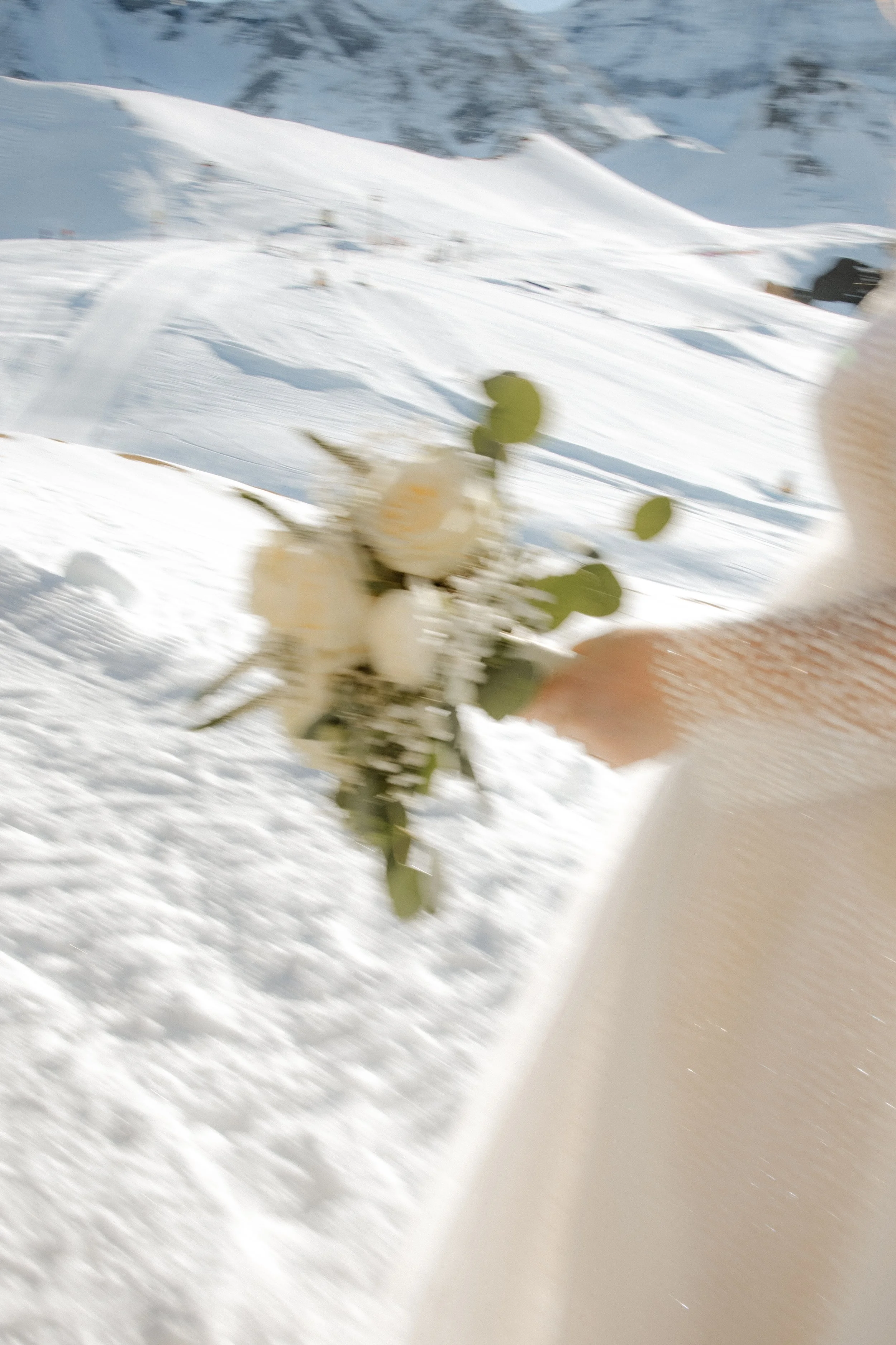 Schneebedeckte Berge und Pisten im Tal, unscharfe weiße Blumen in der Hand einer Person im Vordergrund, auf weißem Kleid. Die Szene ist in den Bergen bei Winterwetter.