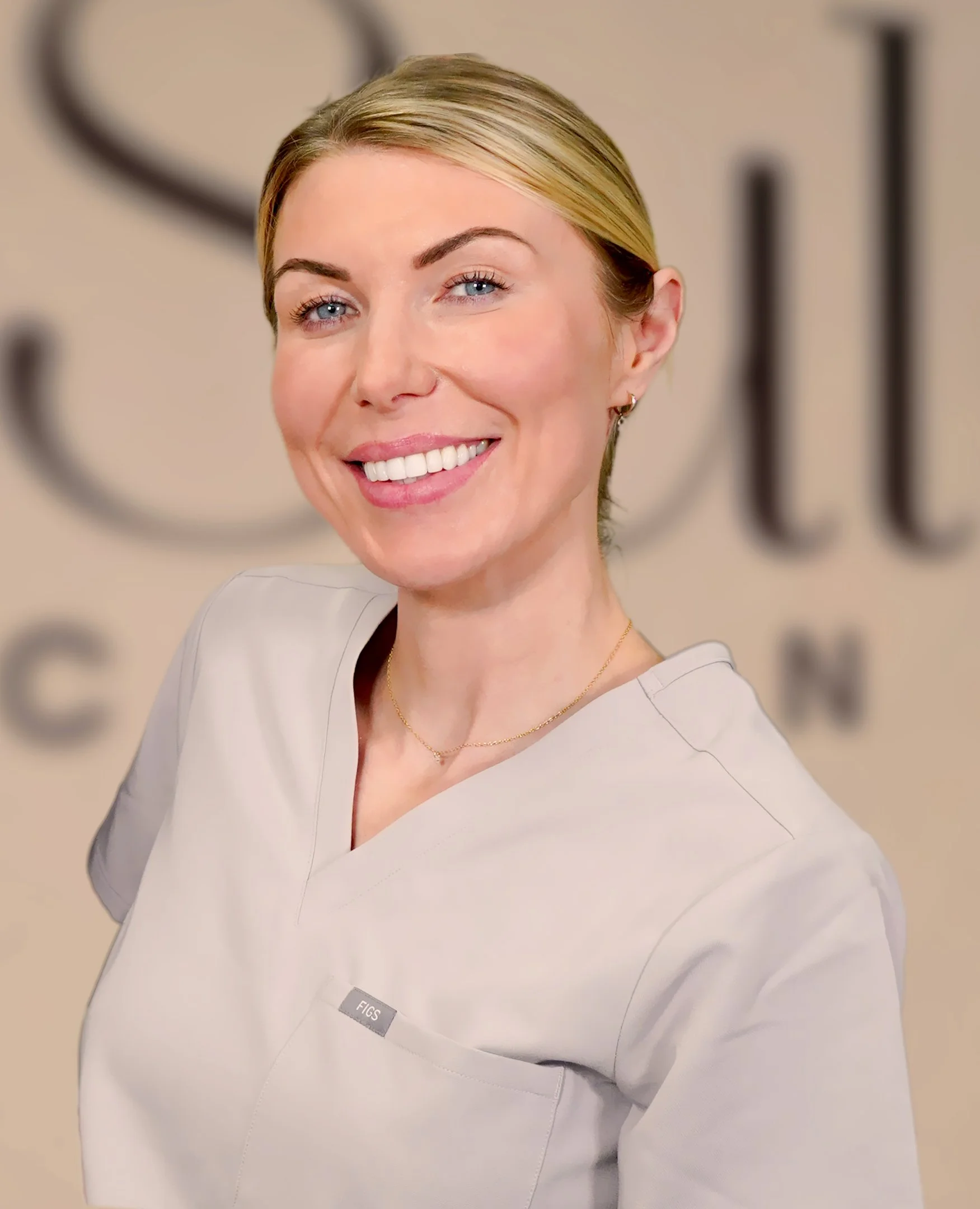 Emma Mills Facialist Skin Specialist. Portrait of a smiling woman with short blonde hair wearing a light-colored medical uniform in a professional setting.