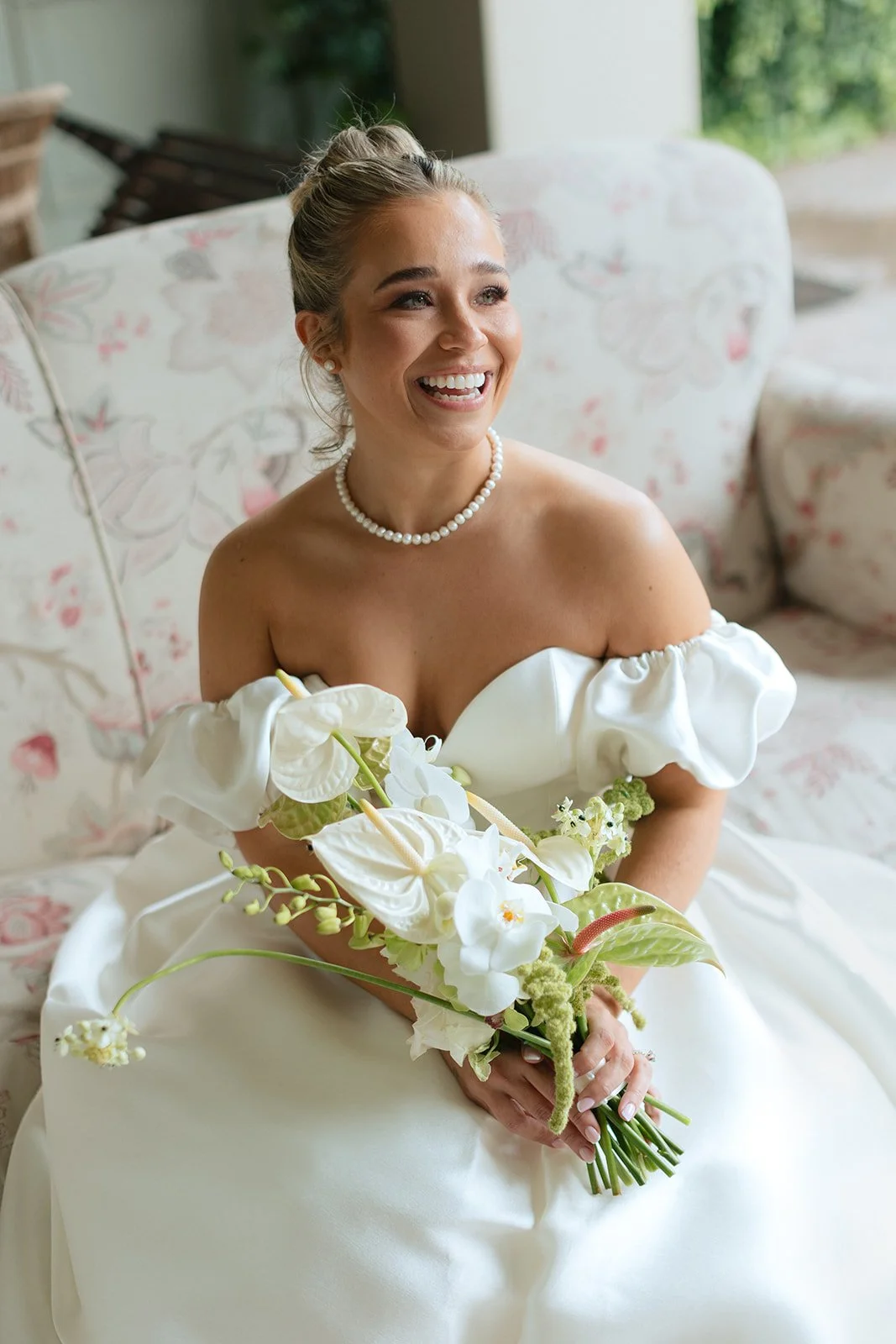 A smiling woman in a white off-shoulder wedding dress with a pearl necklace, sitting on a floral-patterned sofa, holding a bouquet of white flowers with green leaves.