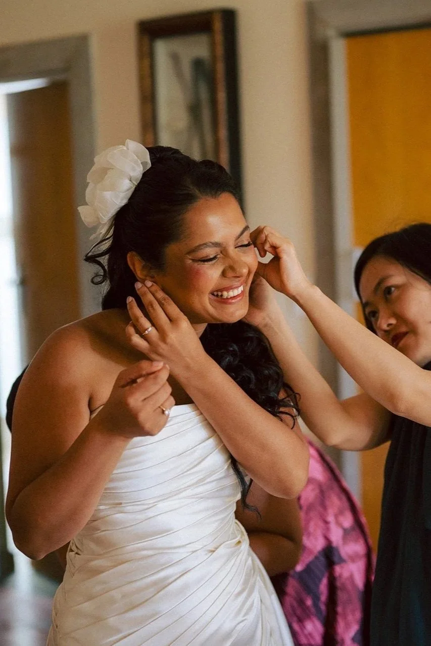 A bride with dark hair and a white strapless wedding gown smiling as someone is adjusting her earring, with another woman smiling nearby.