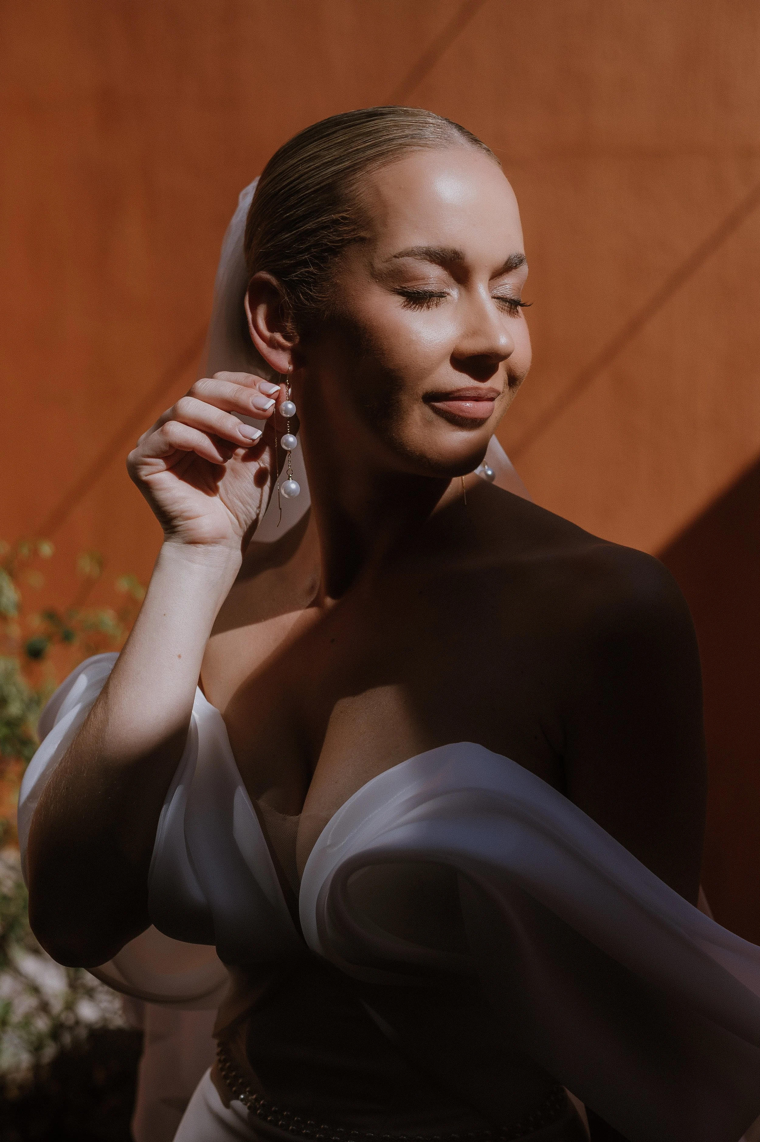 A bride with fair skin and blonde hair styled back, wearing pearl earrings and a white dress off the shoulder, with her eyes closed and a gentle smile, holding her right earring with her right hand, against a warm autumn background.