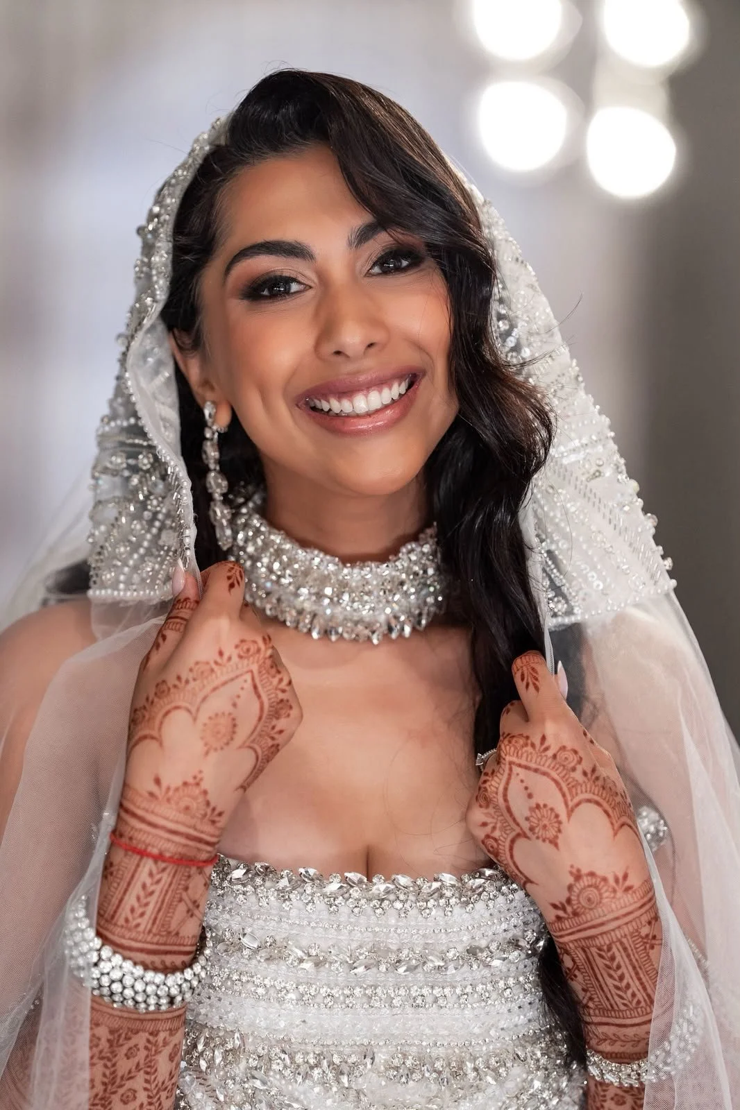 An Indina bride smiling on her wedding day wearing a jeweled head covering, intricate jewelry, and wedding dress, showing detailed henna designs on her hands.