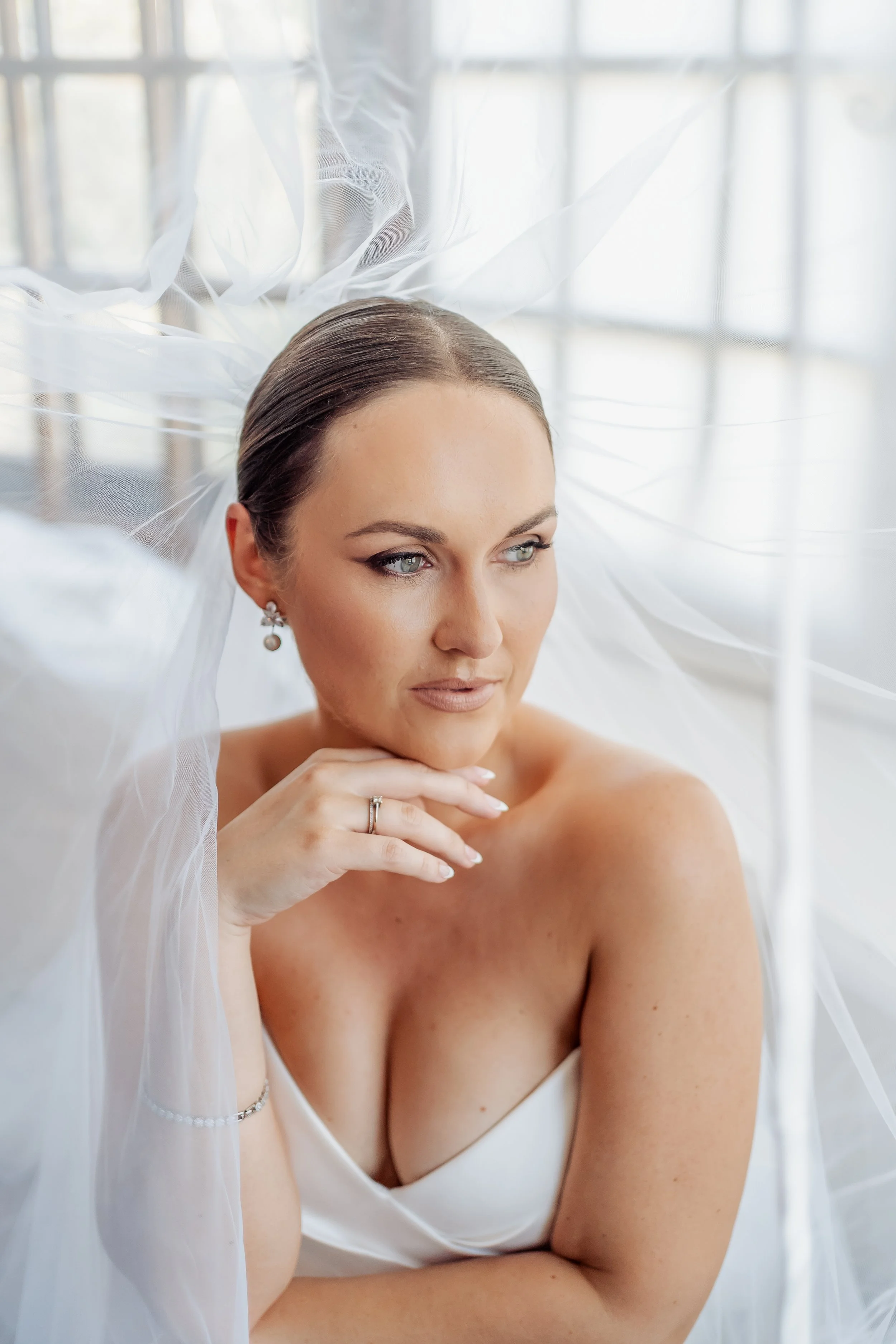A woman with blue eyes and brown hair styled in an elegant updo, wearing pearl earrings and a white strapless top, posed gracefully beside a large window with sheer white curtains.