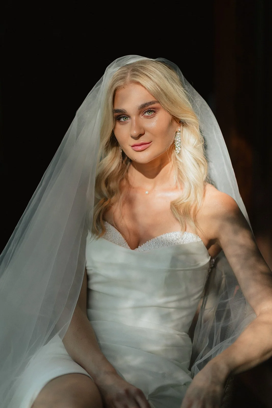 A woman with blonde hair, wearing a white wedding dress and veil, looking at the camera, with a dark background.