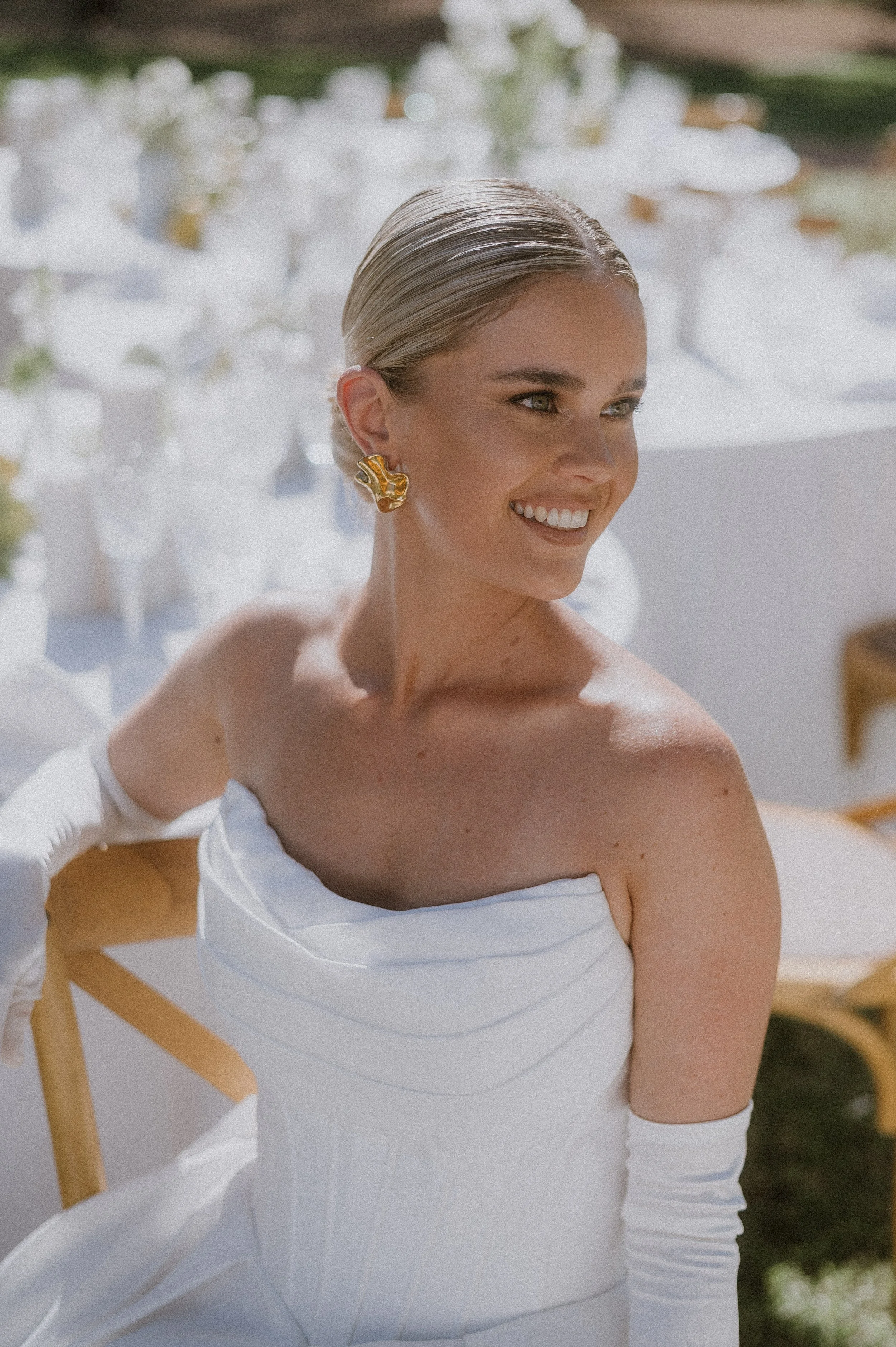 Bride with polished bridal hair and makeup. Ball gown, white gloves and sitting on a wooden chair outdoors.