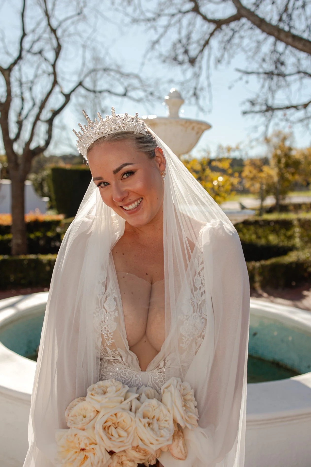 A smiling bride with a tiara and veil holding a bouquet of cream-colored roses outdoors near a fountain and trees with autumn leaves.