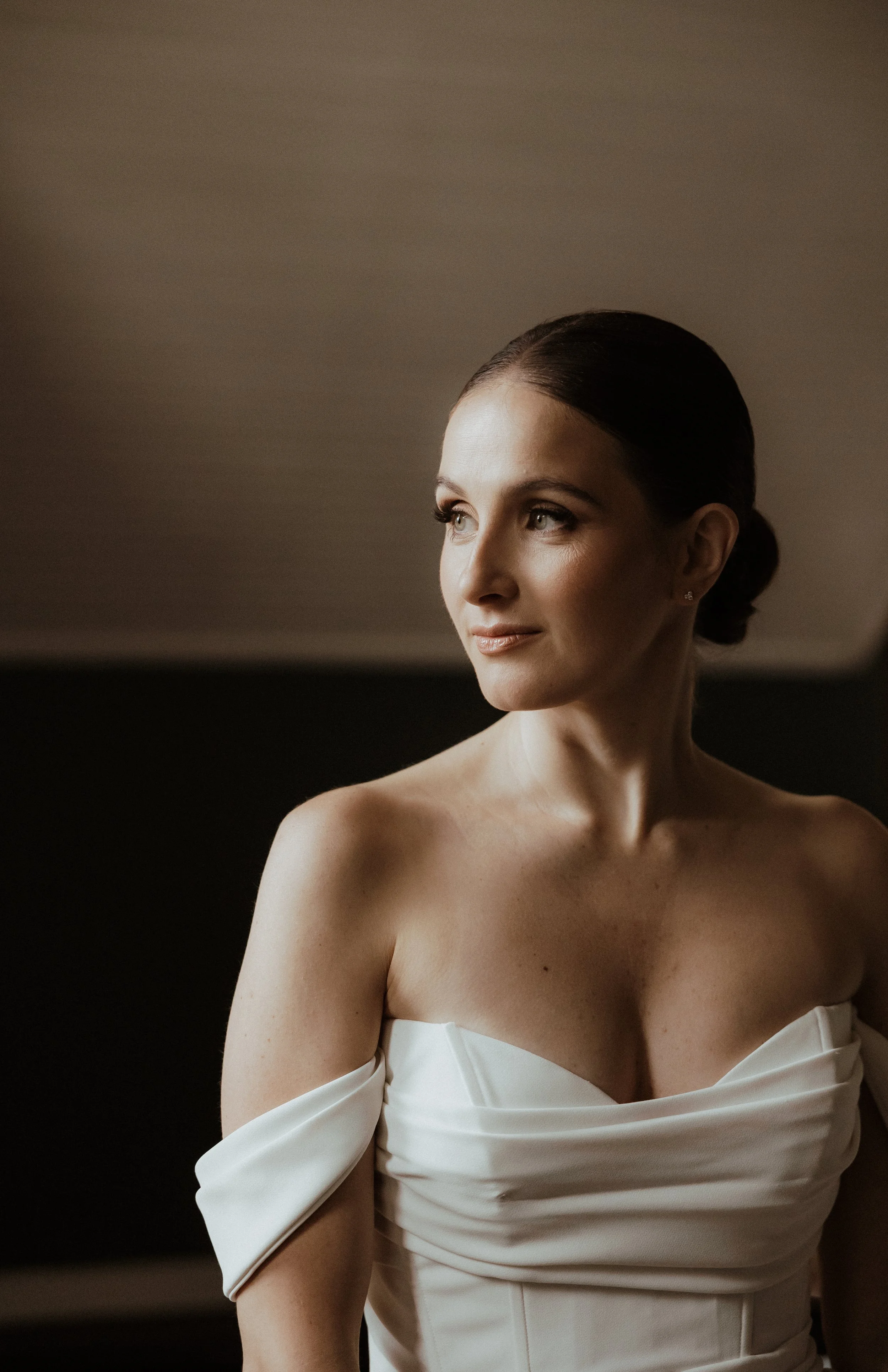 A woman with dark hair styled in a sleek bun, wearing an off-the-shoulder white gown with a draped bodice, looking to her right with a serene expression.