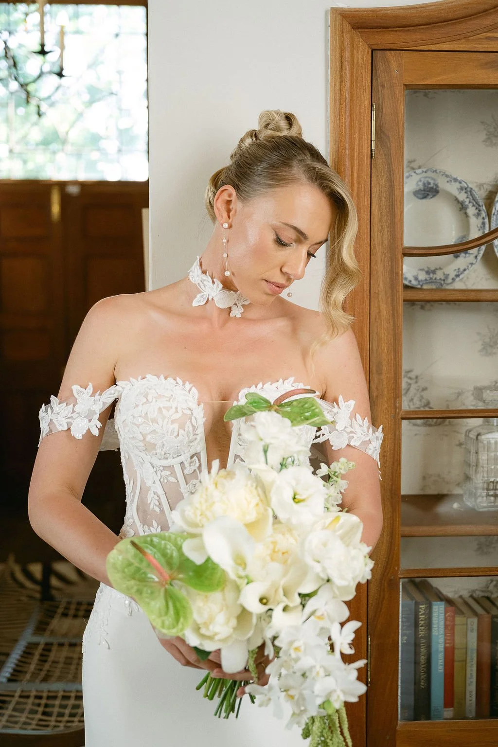 A bride in a white wedding dress holding a bouquet of white flowers, standing indoors next to a wooden cabinet.