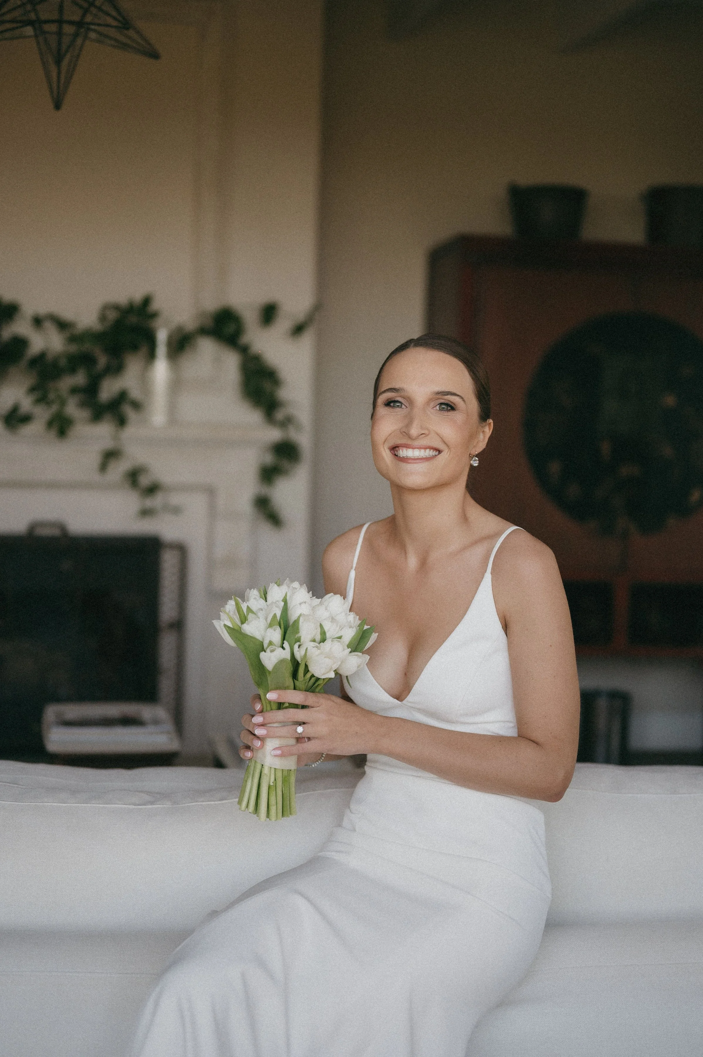 A smiling woman in a white dress sitting on a white surface, holding a bouquet of white flowers, in an indoor setting with plants and a brick fireplace in the background.