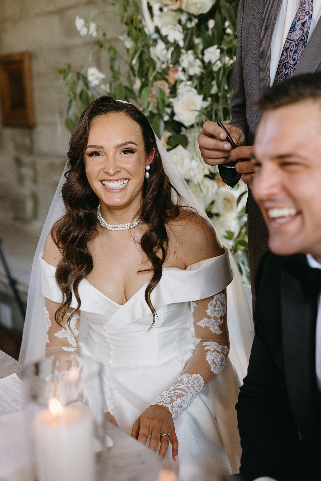 A bride with long dark curly hair wearing a white off-shoulder wedding dress with lace sleeves, a pearl necklace, and pearl earrings, smiling at a wedding ceremony. Retouched for her makeup and hair, behind a floral backdrop with white roses and gree